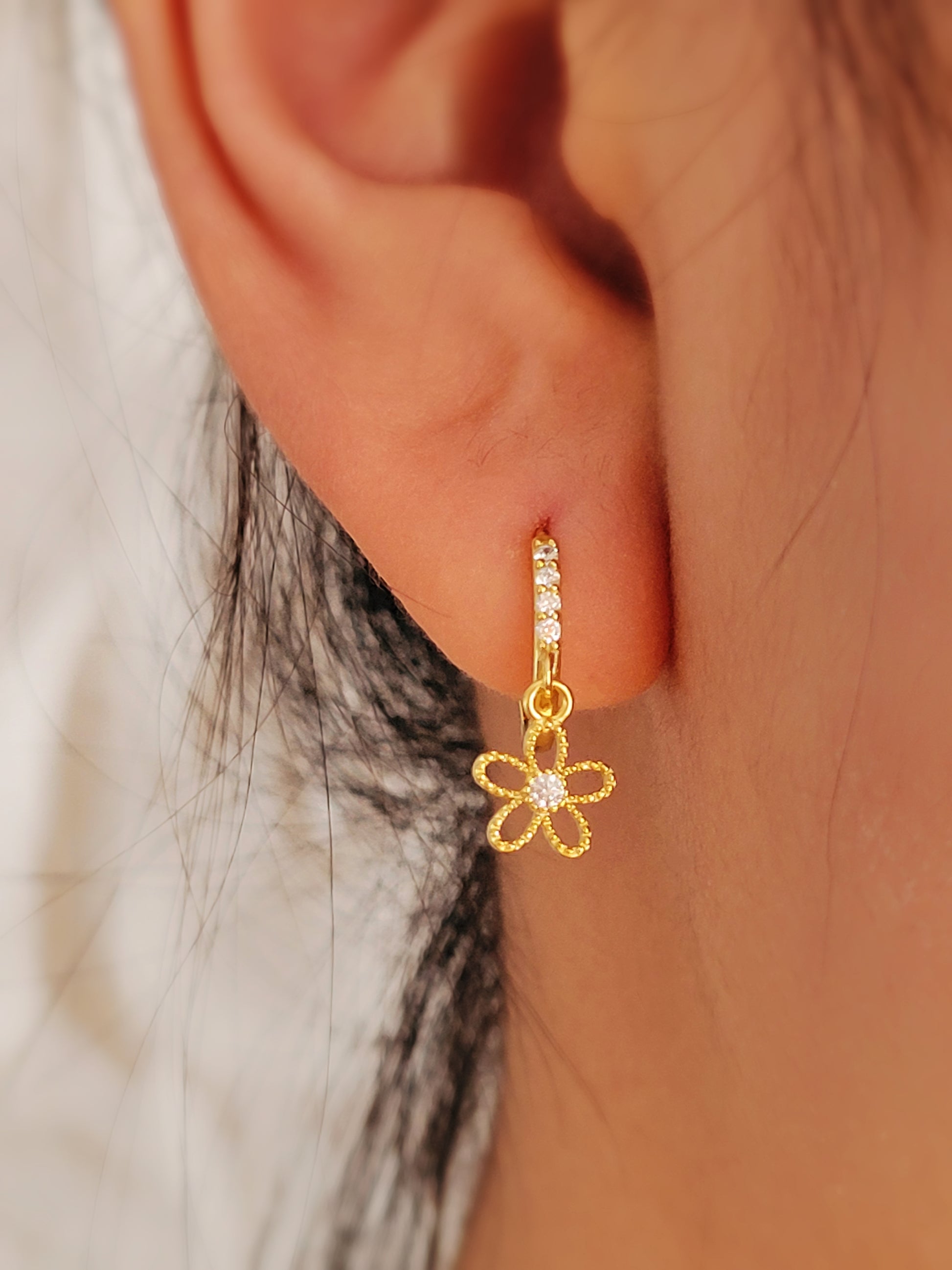 A close-up image of a woman's ear wearing a gold plated huggie earring with a small dangling flower design.