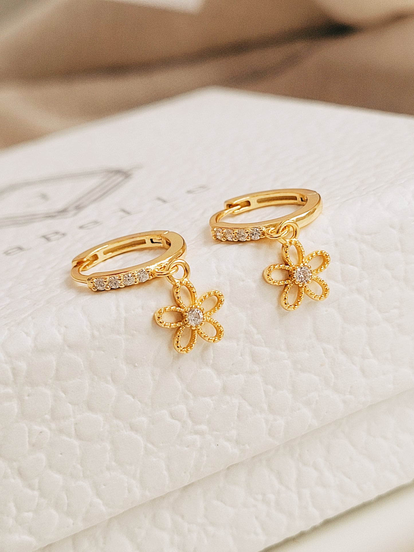 A pair of dainty gold huggie earrings featuring dangly flower charm displayed on a jewellery box. 