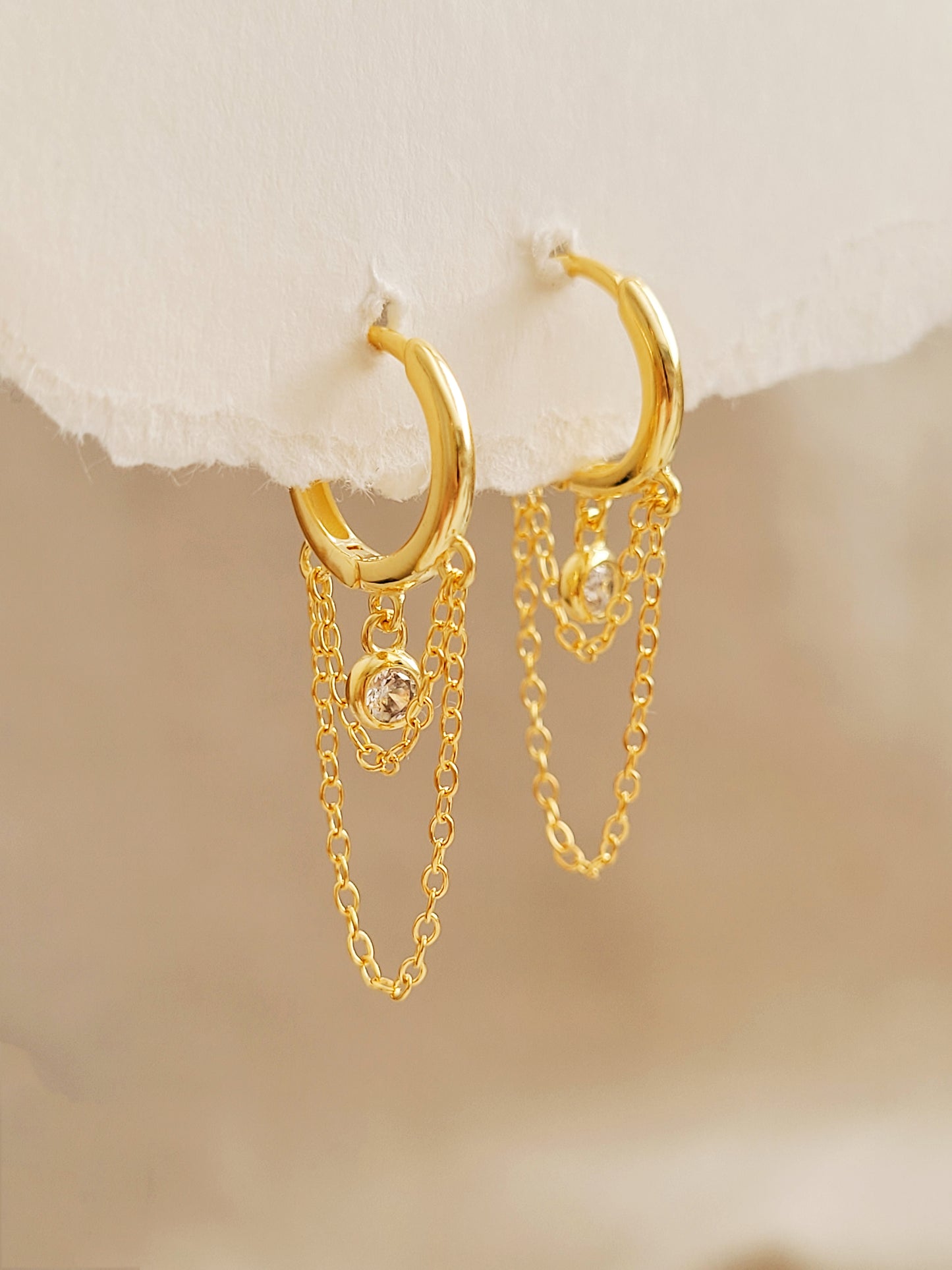 Close-up of a pair of gold double chain huggie earrings with a CZ diamond drop. Crafted in gold plated sterling silver, boho chic summer look.
