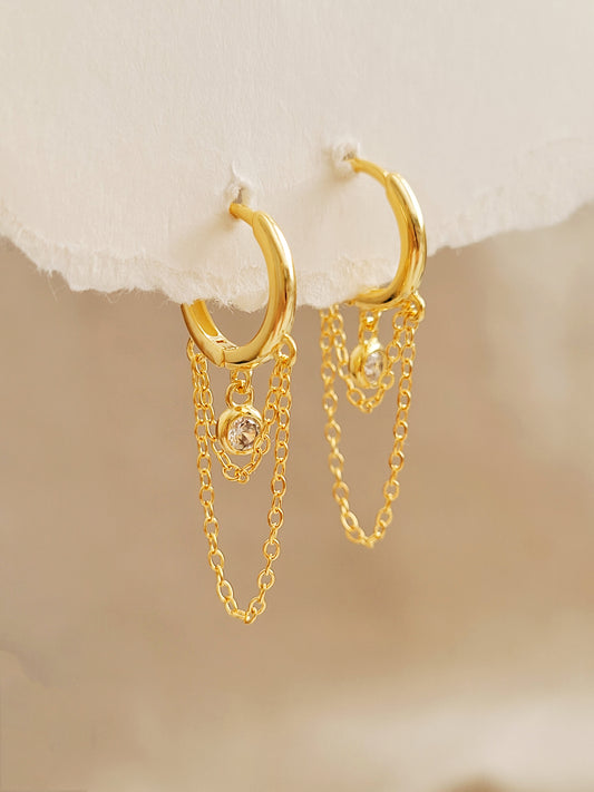 Close-up of a pair of gold double chain huggie earrings with a CZ diamond drop. Crafted in gold plated sterling silver, boho chic summer look.