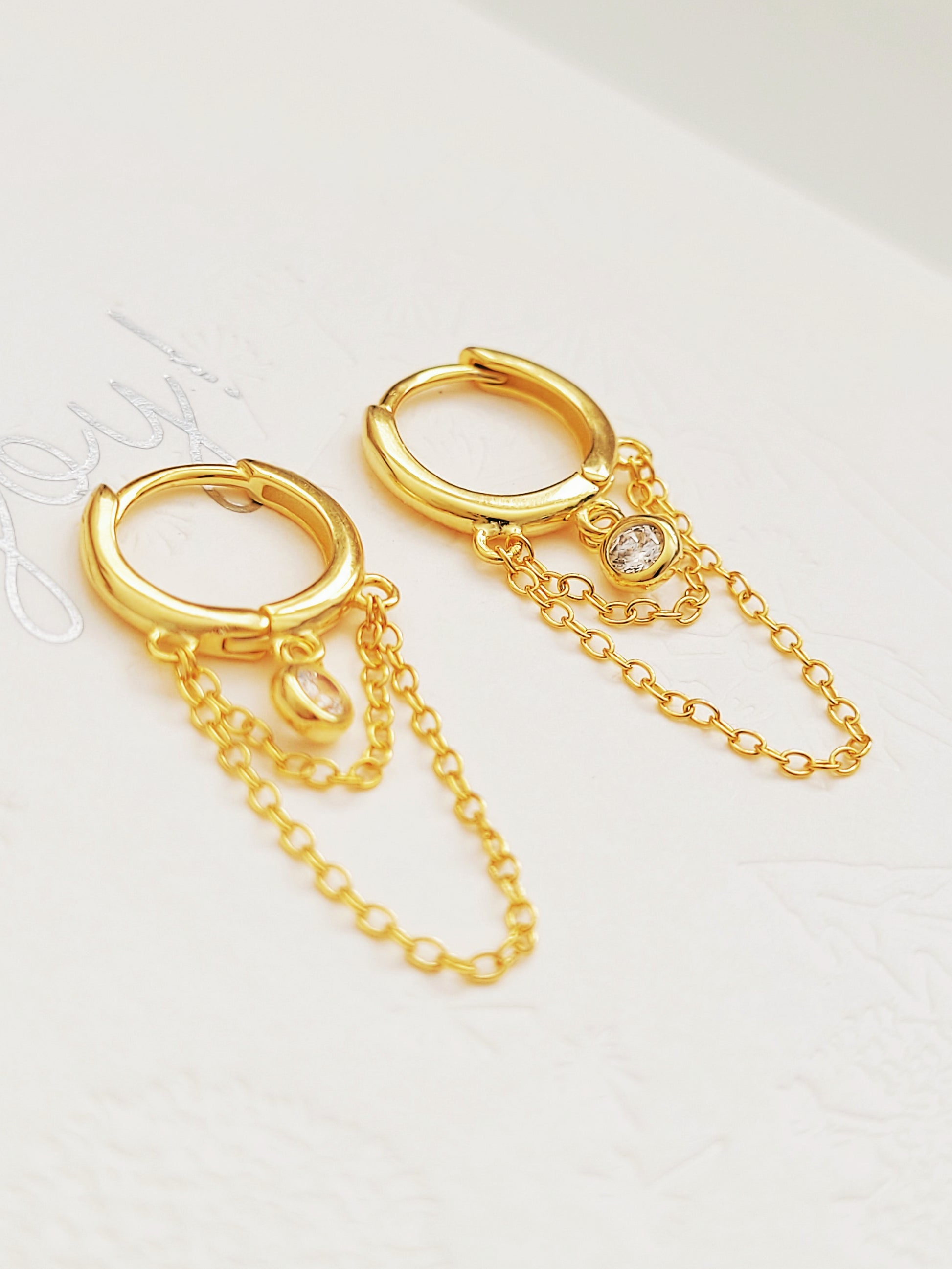 Close-up of a pair of gold double chain huggie earrings with a CZ diamond drop. Crafted in gold plated sterling silver, boho chic summer look.