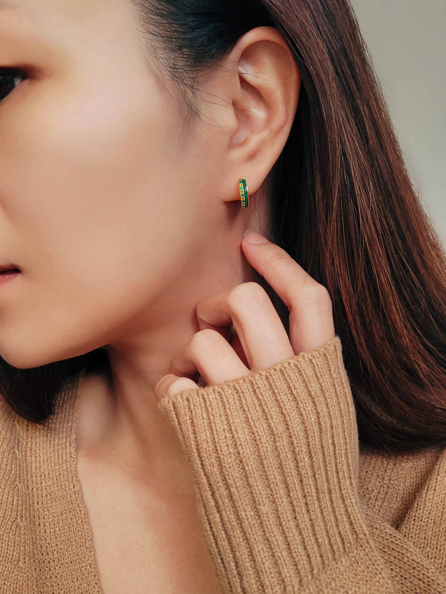 Woman wearing small gold hoop earrings with vibrant green enamel, accents with green and gold crystals.