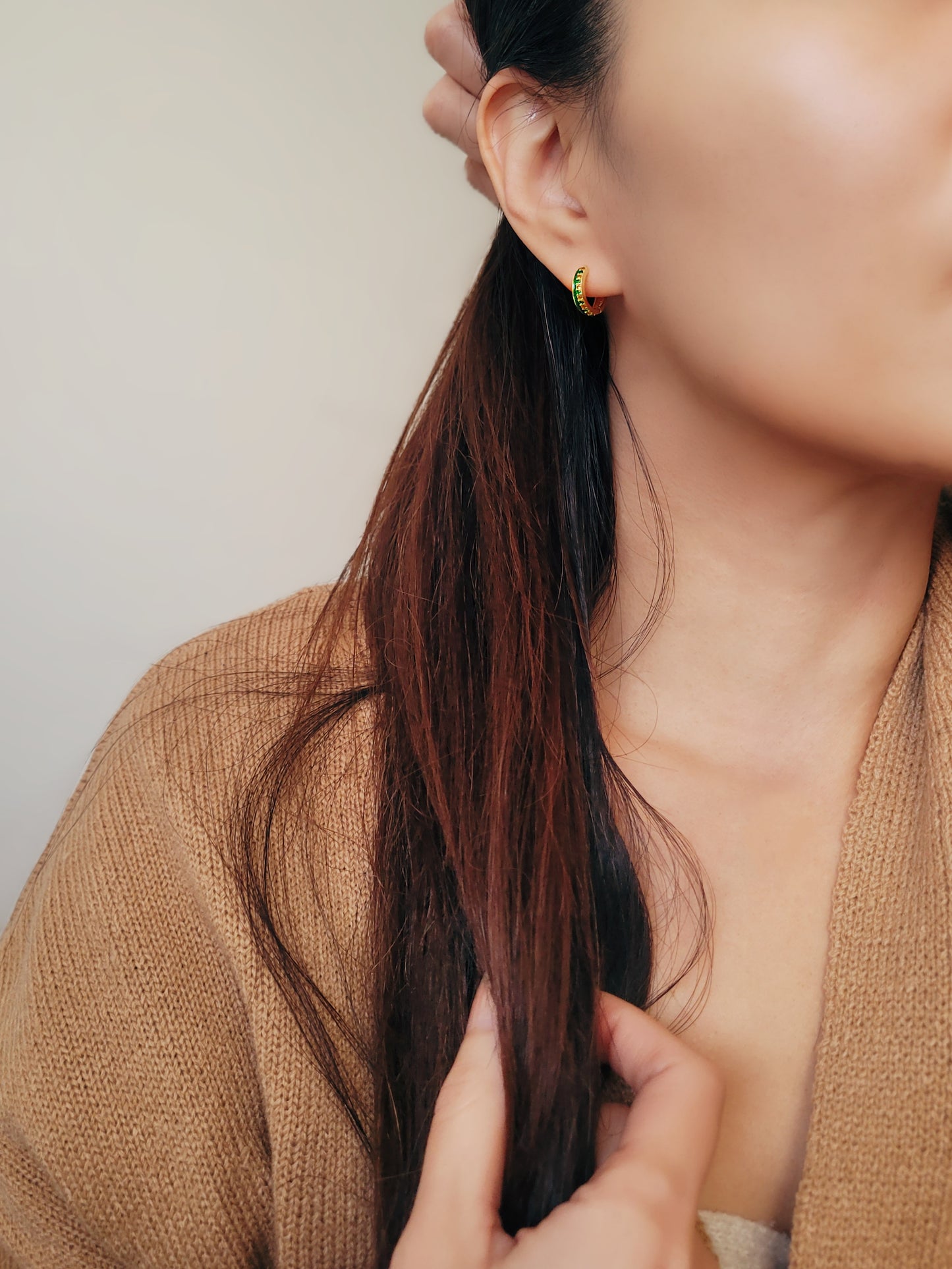 Woman wearing small gold hoop earrings with vibrant green enamel, accents with green and gold crystals.