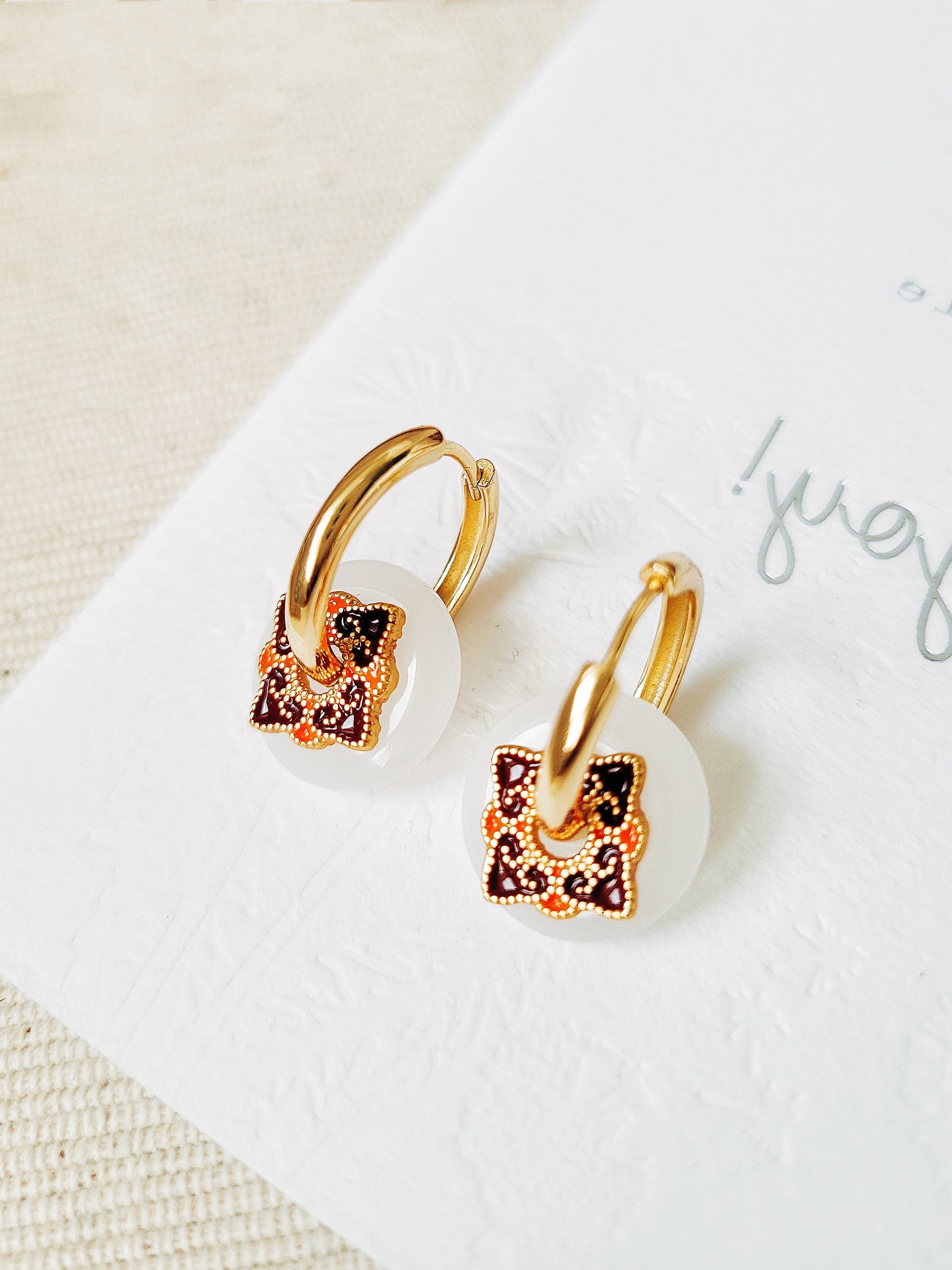 Close-up of enamel hoop earrings with white dangle and vintage tile-inspired charm, set against a neutral background.