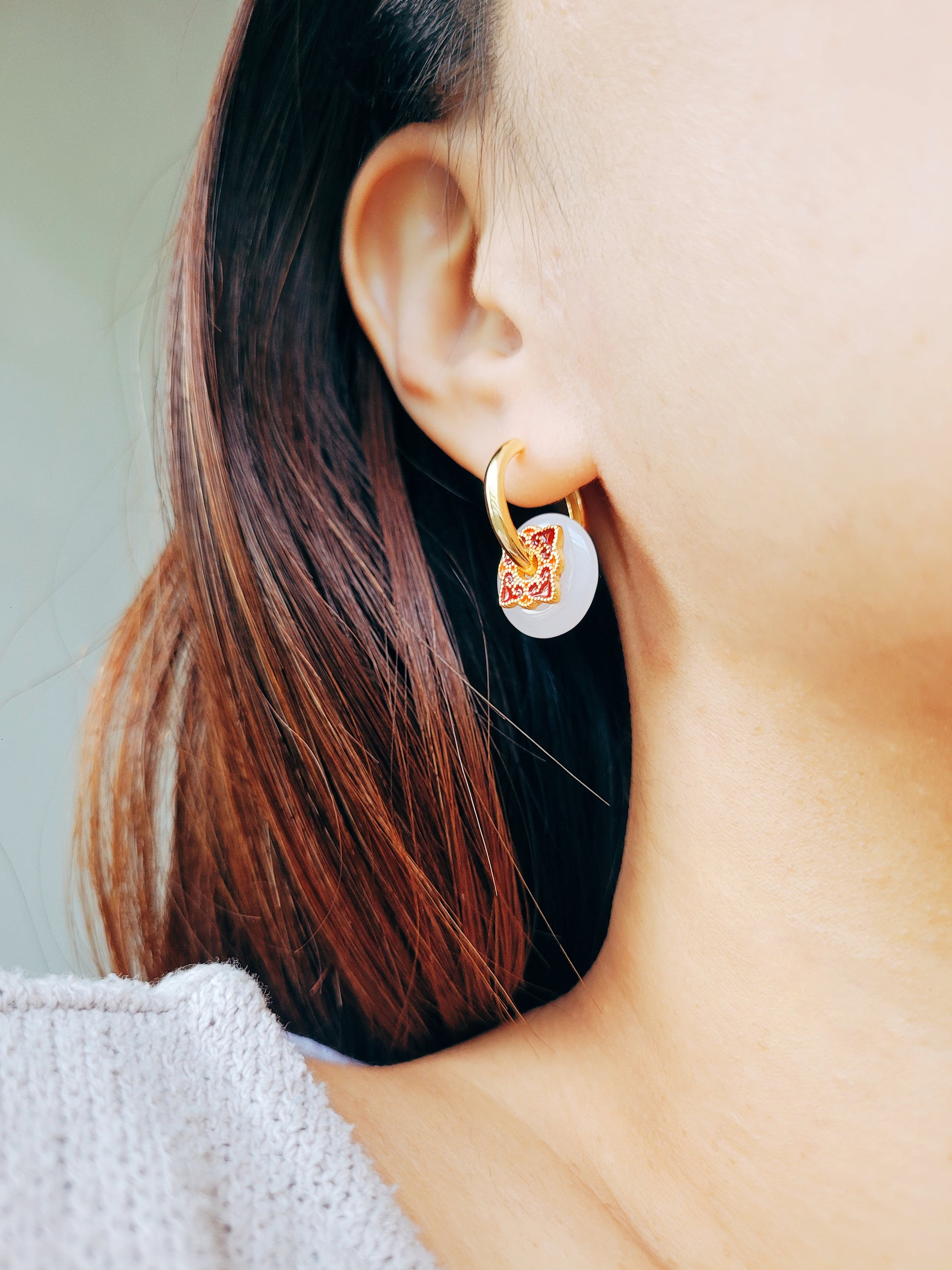 Close up of a woman’s ear wearing an enamel earrings with gold hoops and white dangle.