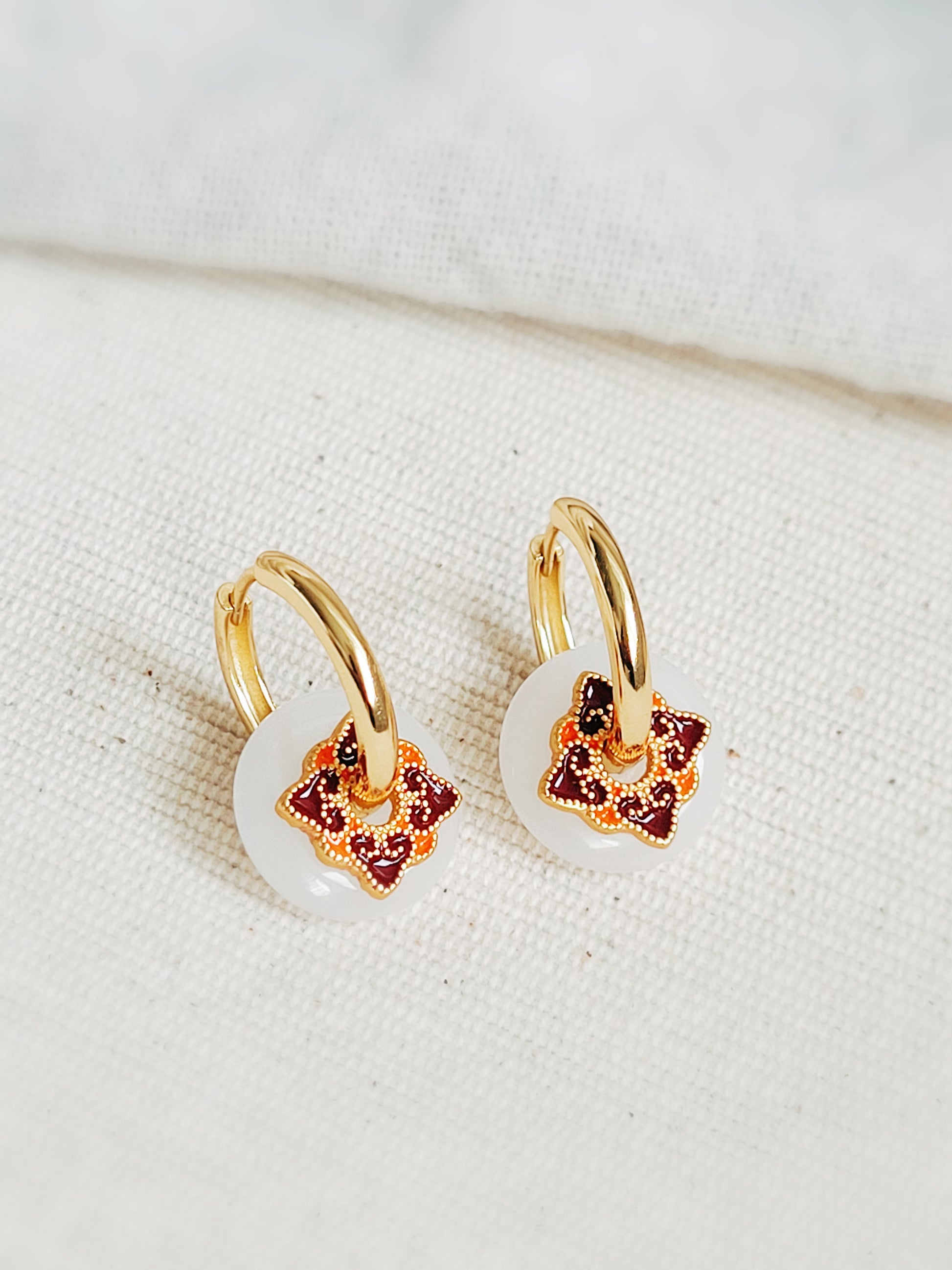 Close-up of enamel hoop earrings with white dangle and vintage tile-inspired charm, set against a neutral background.