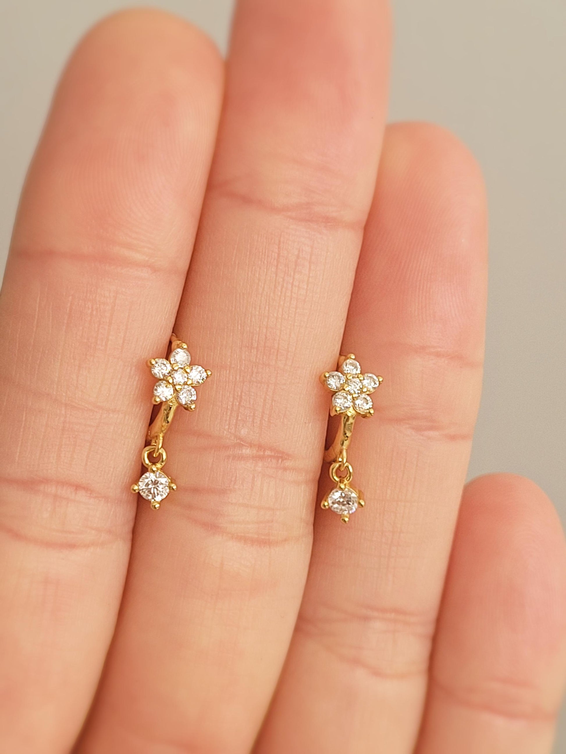 Gold flower earrings with small diamonds held between fingers against a neutral background