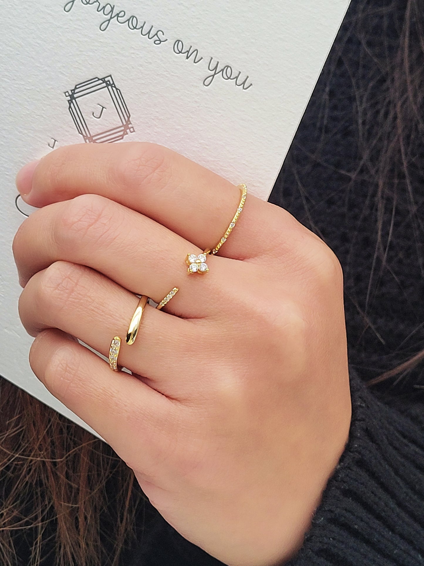 A hand wearing three elegant stacking rings, each featuring gold accents. One ring is open-ended with a sleek, minimalistic design, another is encrusted with small sparkling stones, and the third ring has a delicate clover-shaped cluster of gemstones. 