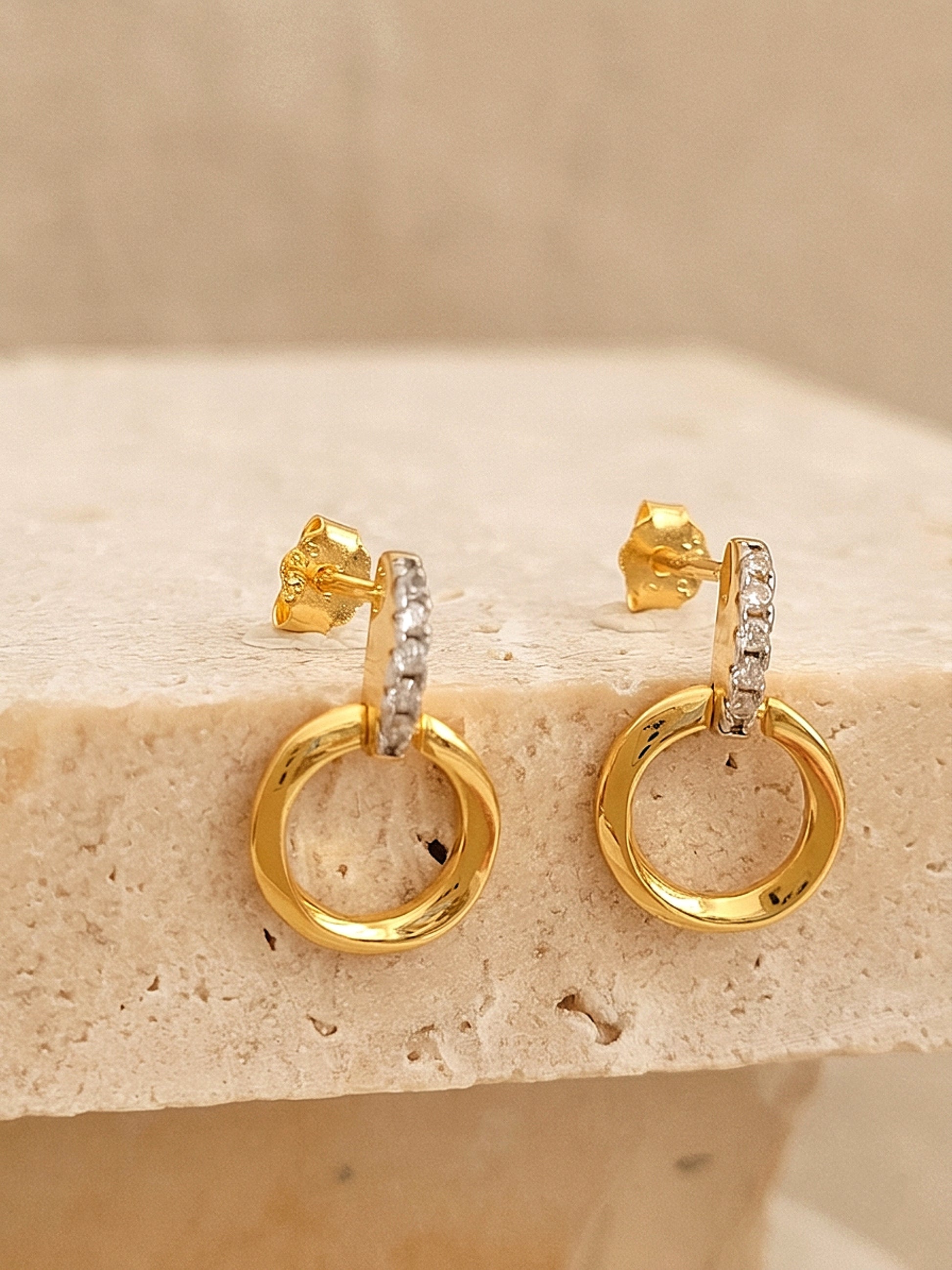 A pair of halo drop stud earrings in gold vermeil with cz diamond accents displayed on an esthetic beige color stone surface
