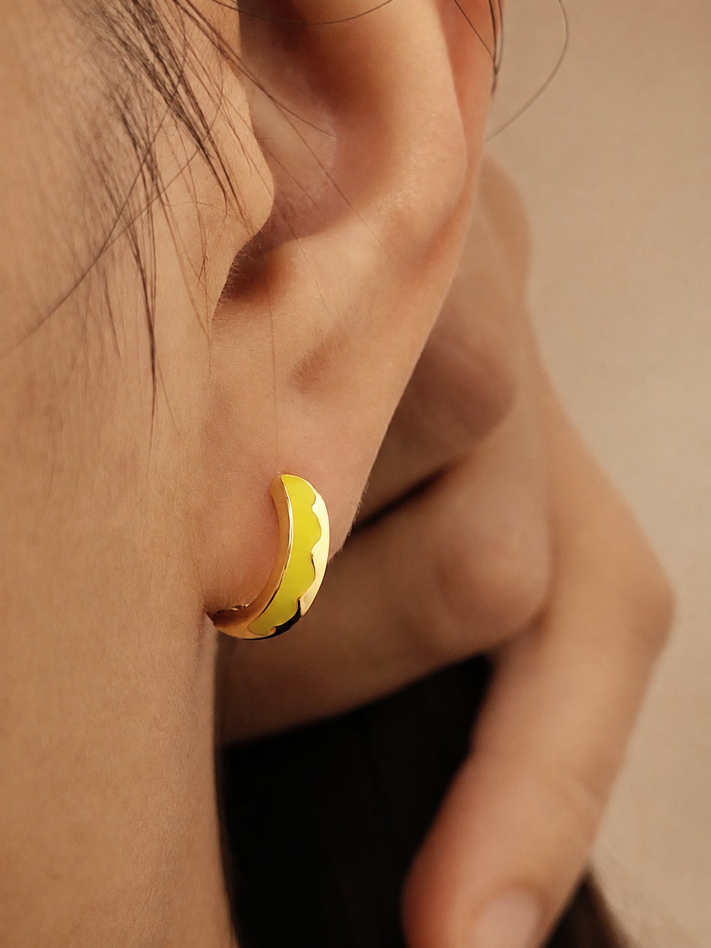 A gold-coloured hoop earring with a yellow enamel design. The earring is a small, curved shape, with a bright yellow colour on the outer edge and a black accent. The earring is worn on the ear.