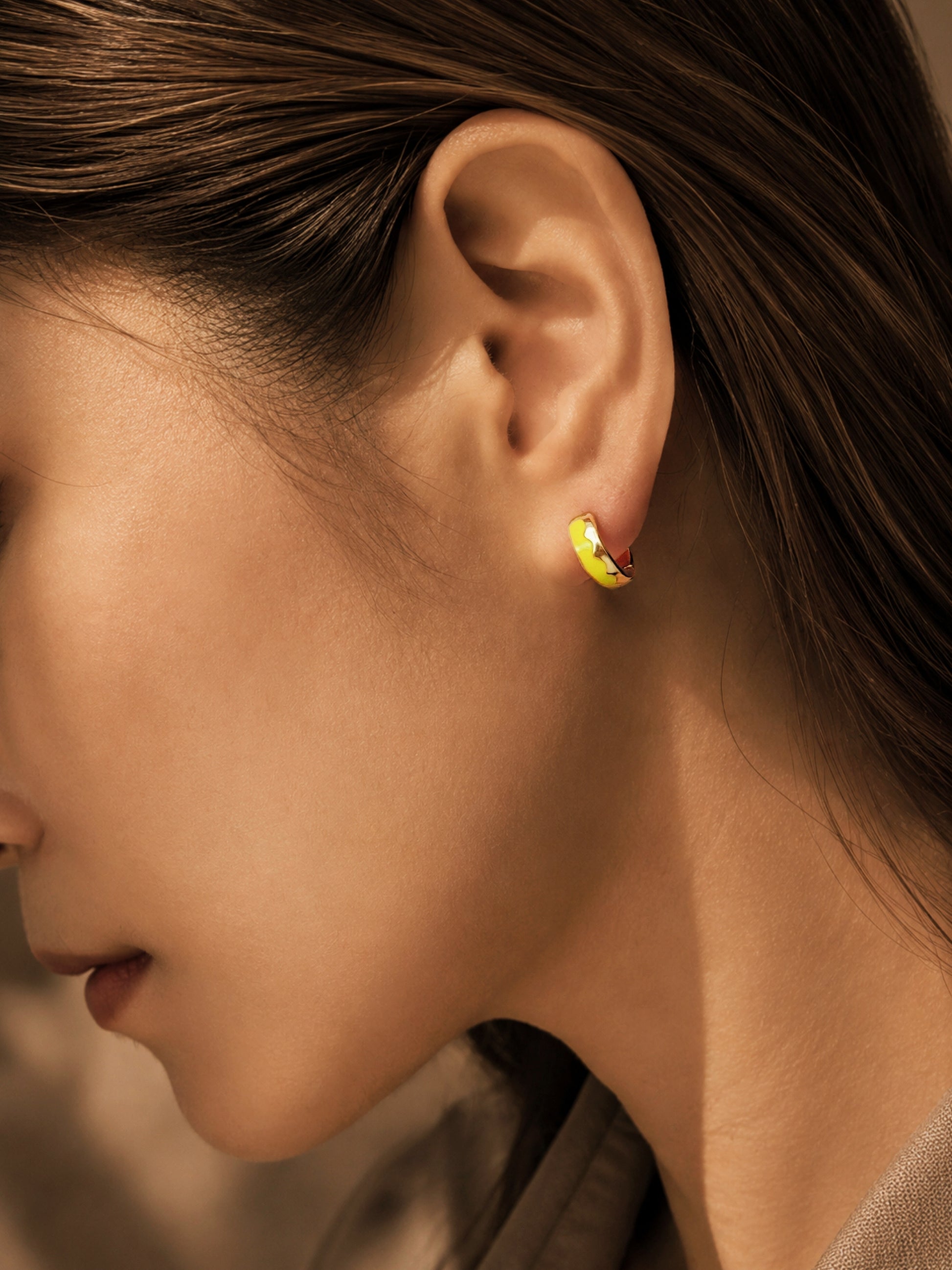 A close-up of an ear with a small gold hoop earring. The earring has a bright wave pattern yellow enamel with simple, modern design, suitable for everyday wear.