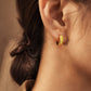 Close-up of a small gold hoop earring with a bright yellow enamel finish. The earring is worn on an ear, showcasing its curved design and vibrant colour. The jewellery is a simple yet stylish accessory.