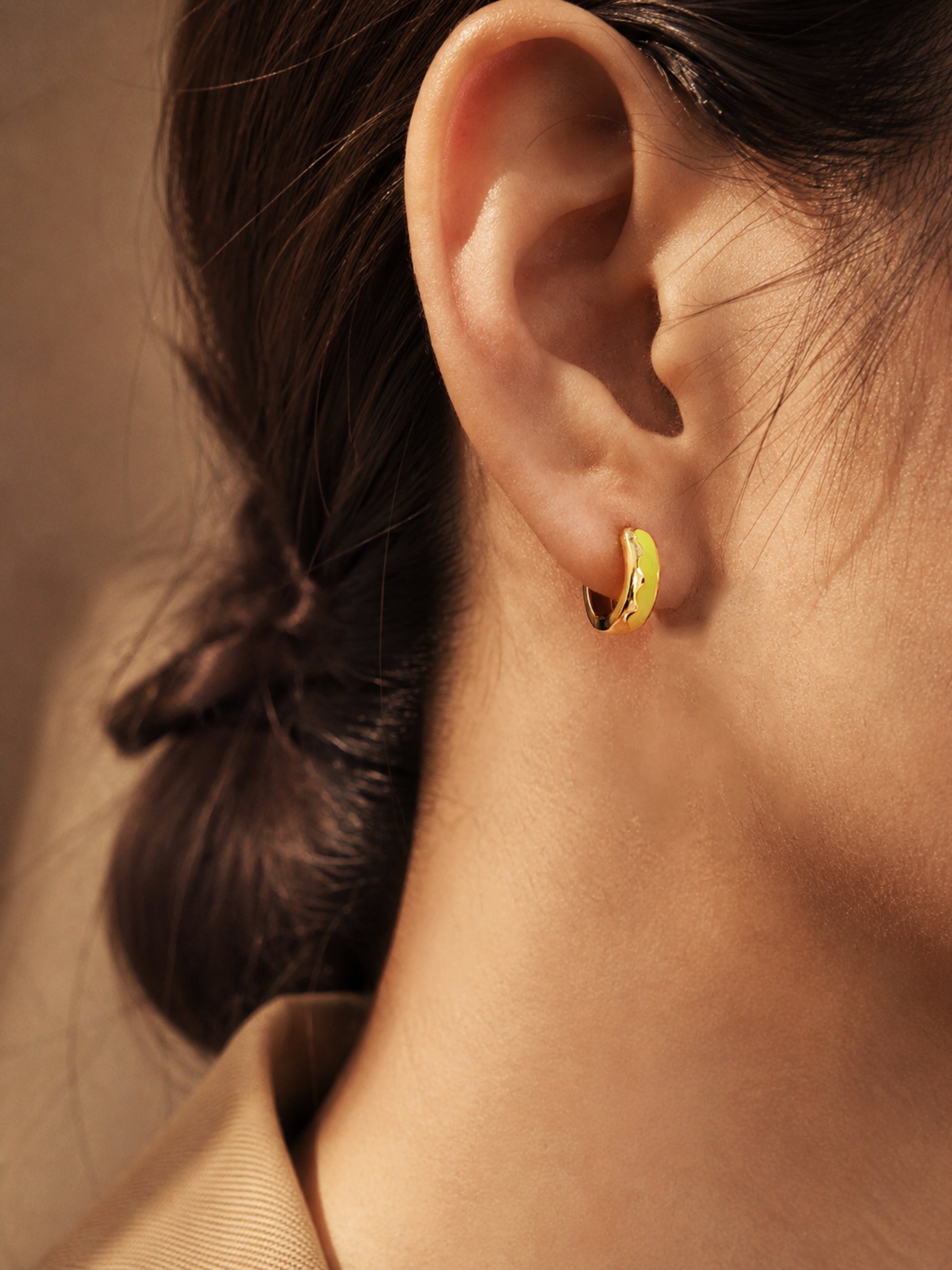 Close-up of a small gold hoop earring with a bright yellow enamel finish. The earring is worn on an ear, showcasing its curved design and vibrant colour. The jewellery is a simple yet stylish accessory.