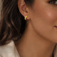 A close-up shot of a woman's ear, showcasing a small, gold hoop earring with a vibrant yellow enamel coating. The earring has a simple, curved shape. The person is wearing a white shirt.