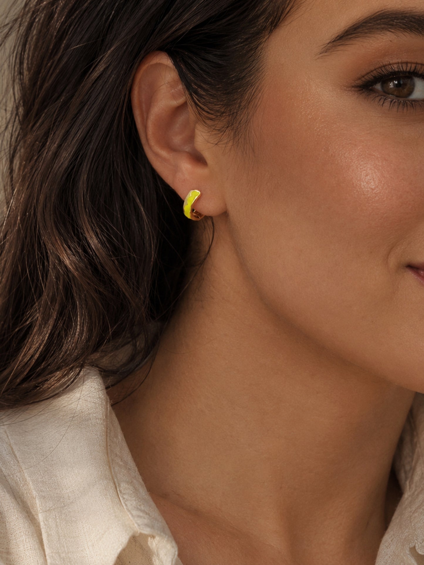 A close-up shot of a woman's ear, showcasing a small, gold hoop earring with a vibrant yellow enamel coating. The earring has a simple, curved shape. The person is wearing a white shirt.