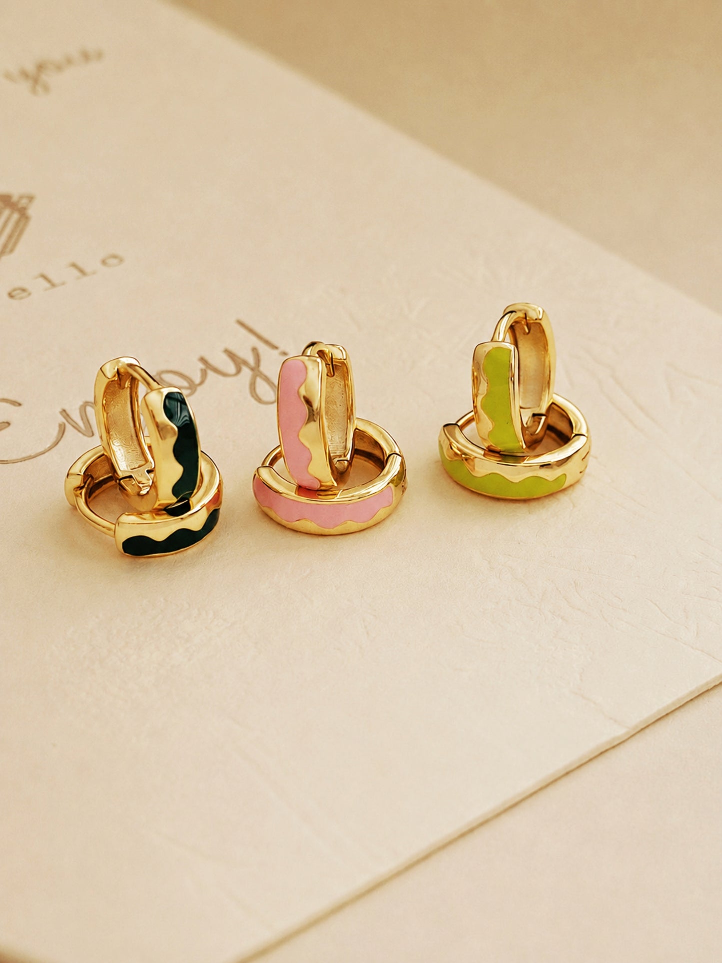 Three pairs of gold-coloured hoop earrings with colourful enamel accents. One pair features black and green enamel, another pink, and the last pair has green and yellow enamel. The earrings are displayed on a light-coloured surface.