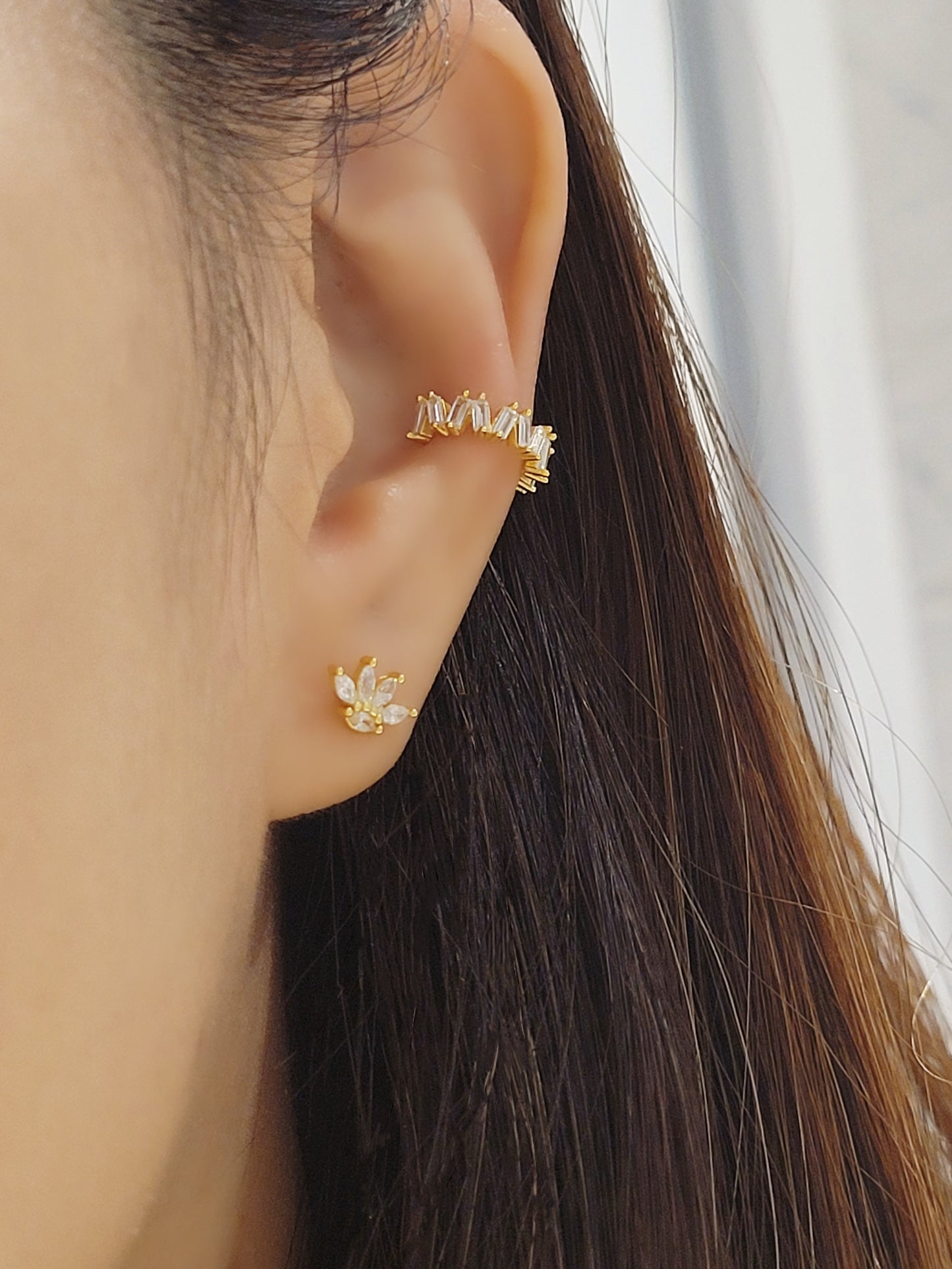 Close-up of a woman's ear wearing a gold vermeil ear cuff with baguette-cut stones, showcasing its modern, geometric design. Paired with a small stud earring in a floral pattern, the cuff sits comfortably on the upper ear without needing a piercing.