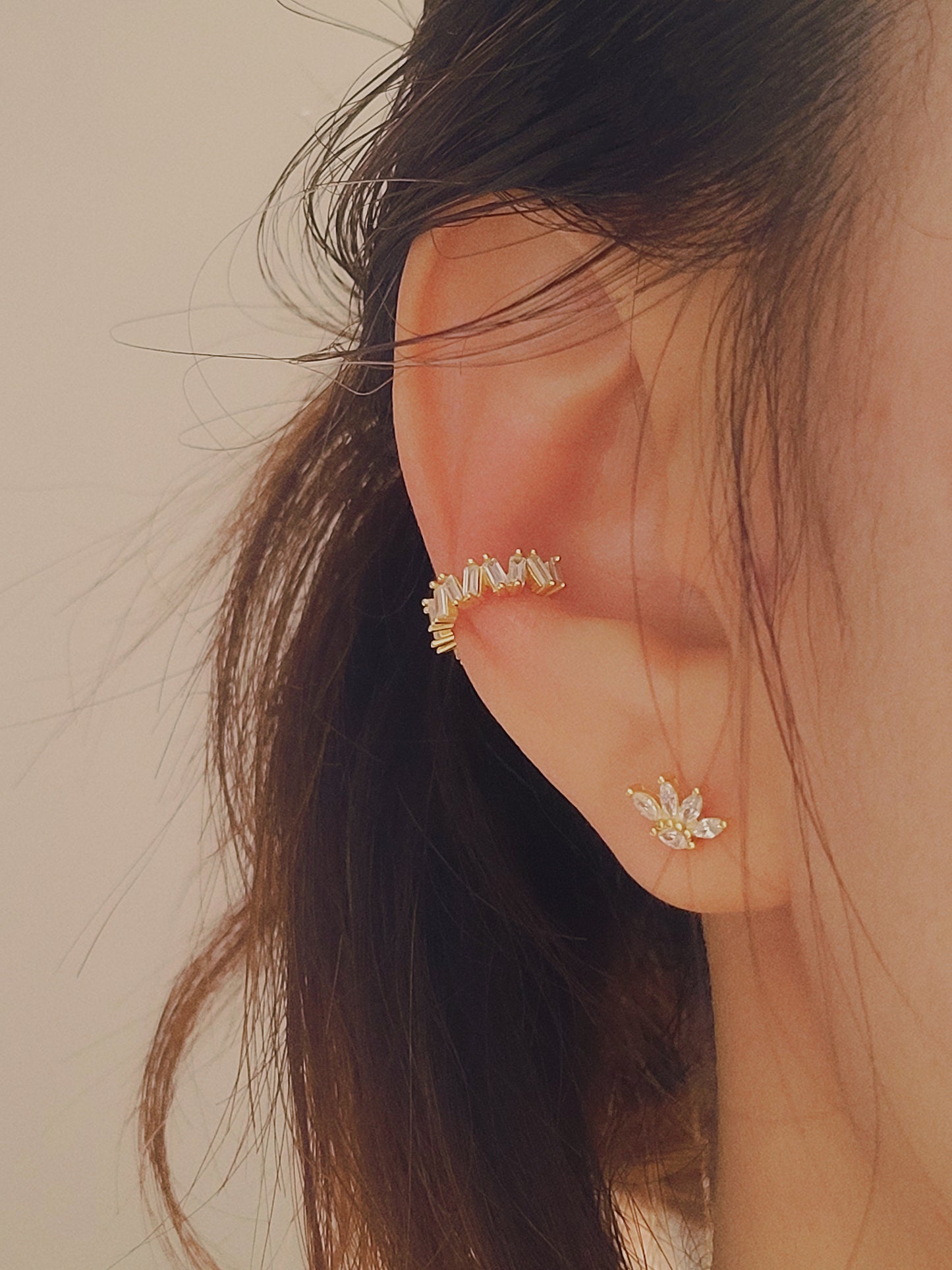 Close-up of a woman's ear wearing a gold vermeil ear cuff with baguette-cut stones, showcasing its modern, geometric design. Paired with a small stud earring in a floral pattern, the cuff sits comfortably on the upper ear without needing a piercing.