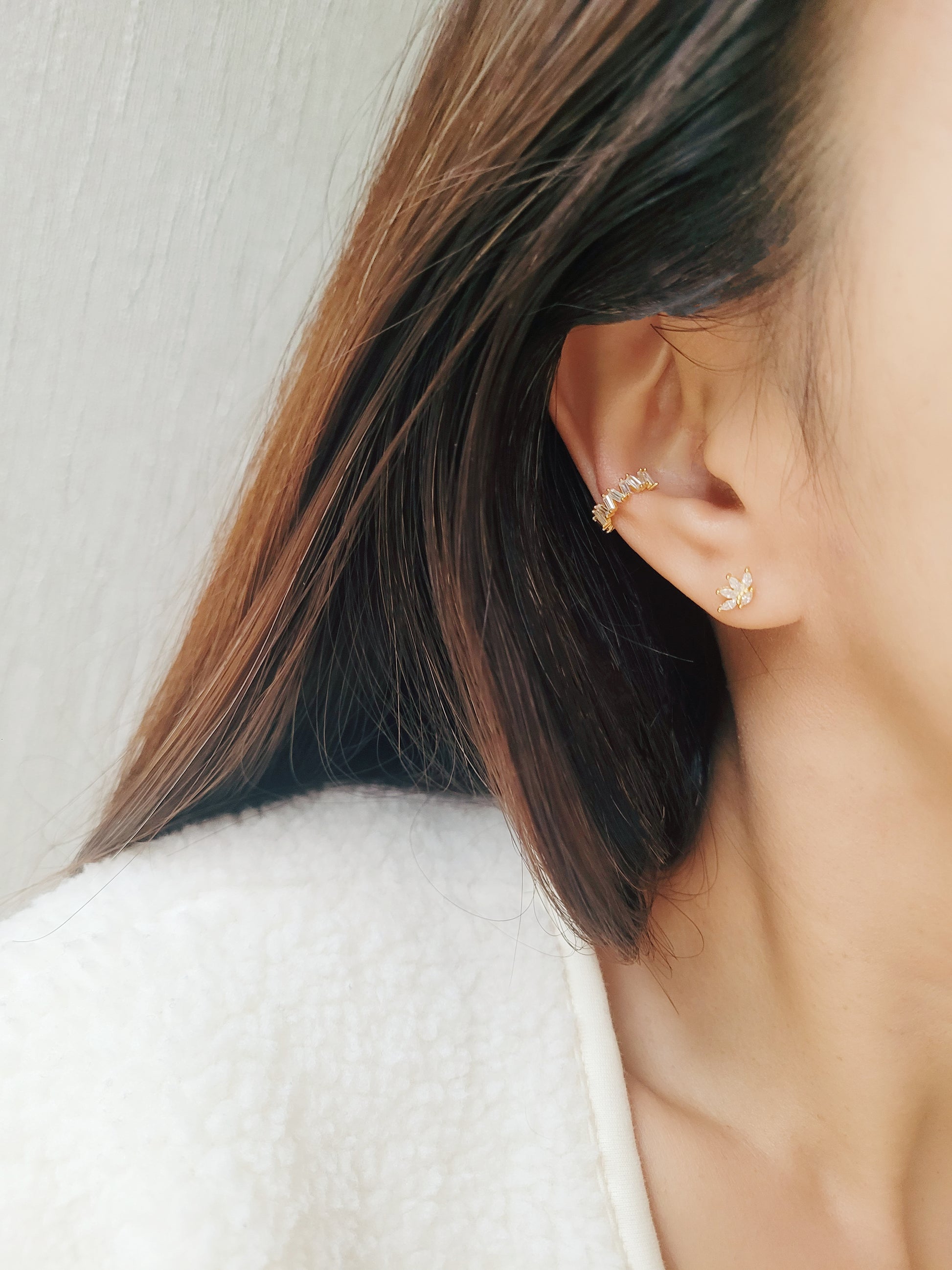 Close-up of a woman's ear wearing a gold vermeil ear cuff with baguette-cut stones, showcasing its modern, geometric design. Paired with a small stud earring in a floral pattern, the cuff sits comfortably on the upper ear without needing a piercing.