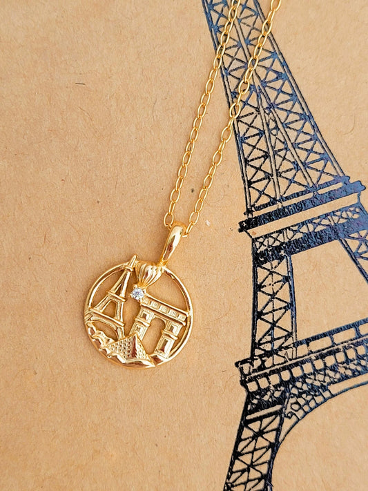 Gold necklace with a Paris-themed pendant on a beige background with a black outline of the Eiffel Tower.