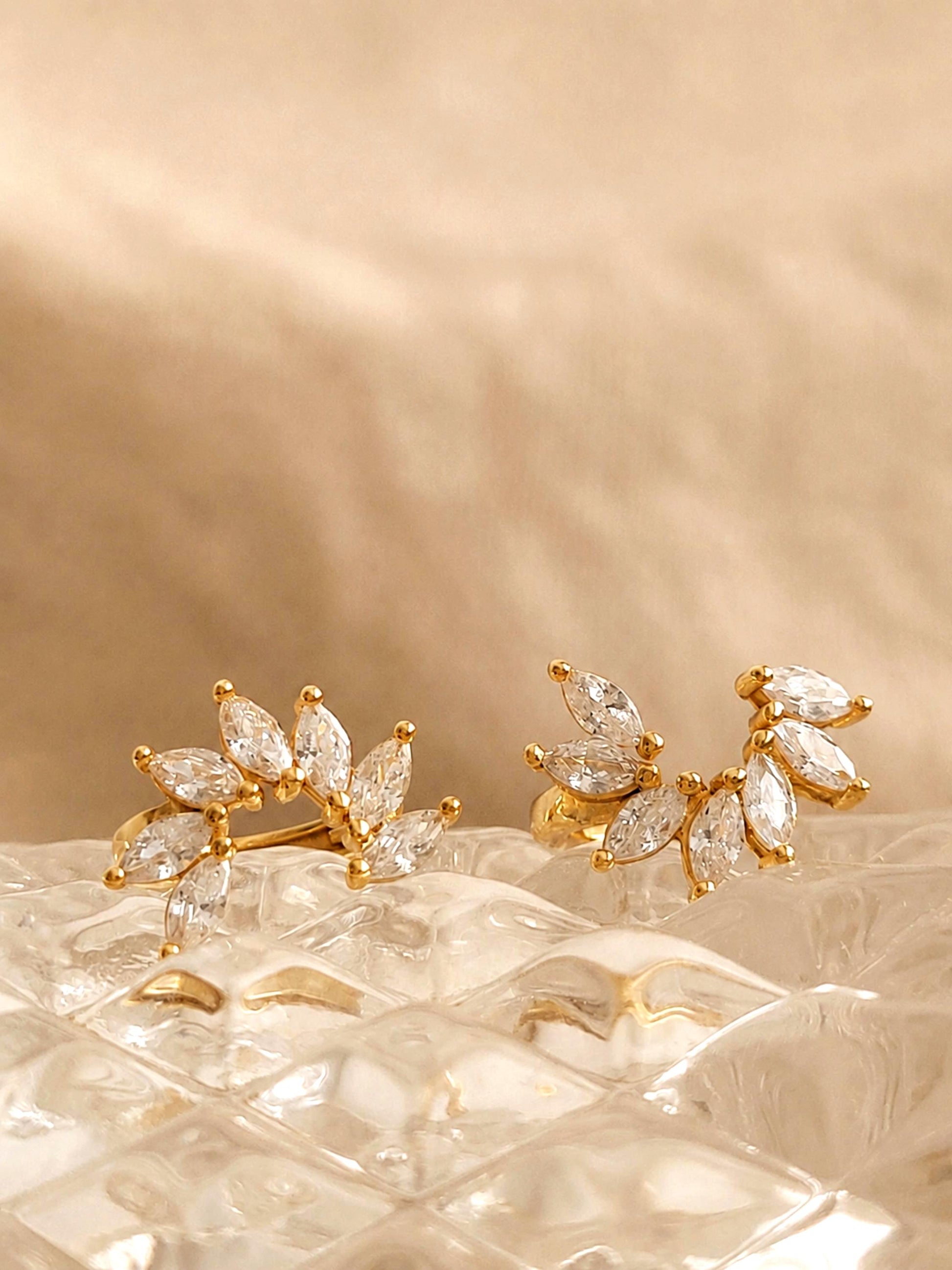 A pair of silver flower huggie earrings featuring floral arrangement shape with cz gemstone, displayed on a luxury beige glass surface.