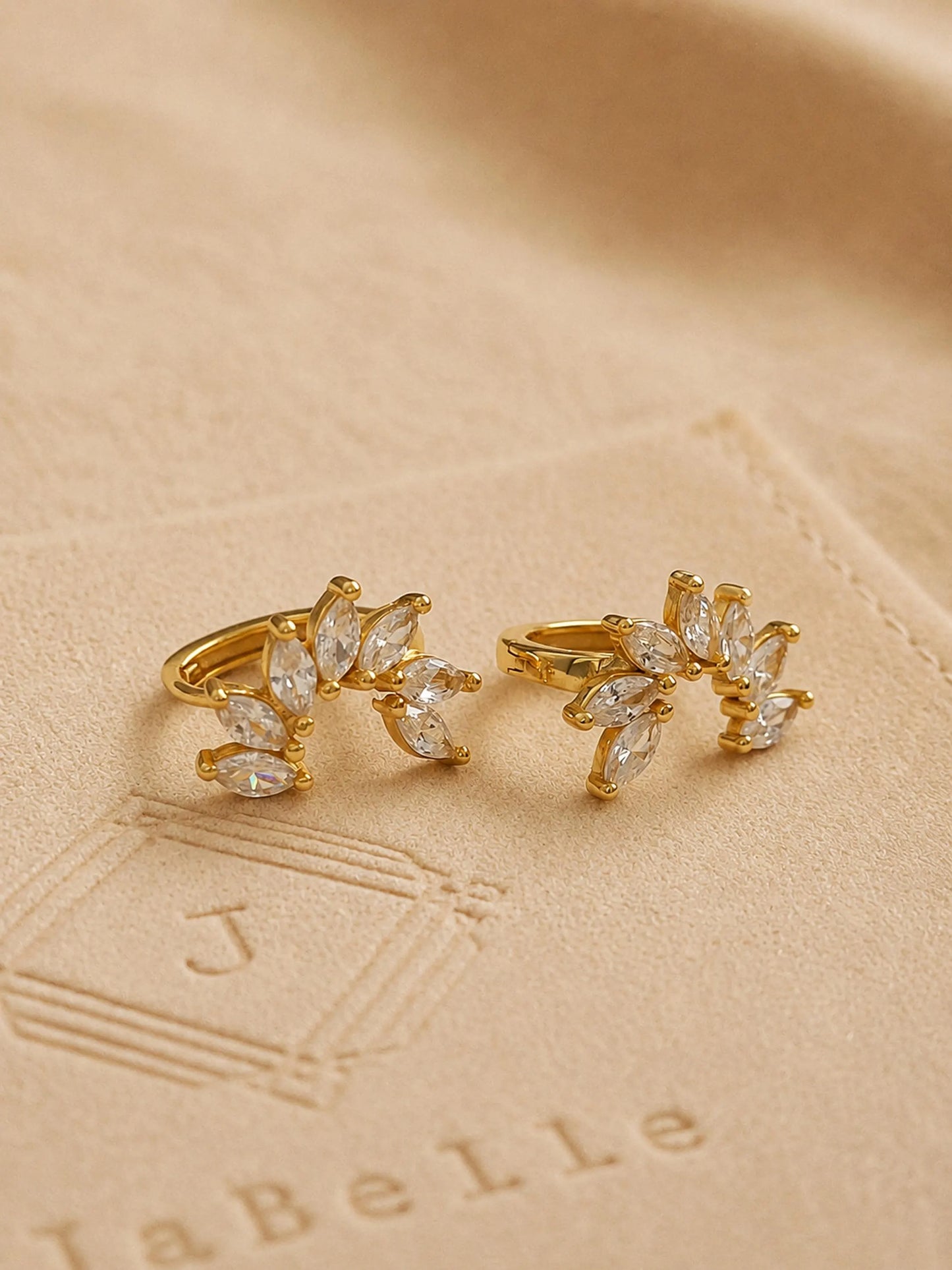 A pair of gold flower huggie earrings featuring floral arrangement shape with cz gemstone, displayed on a luxury beige stone surface.