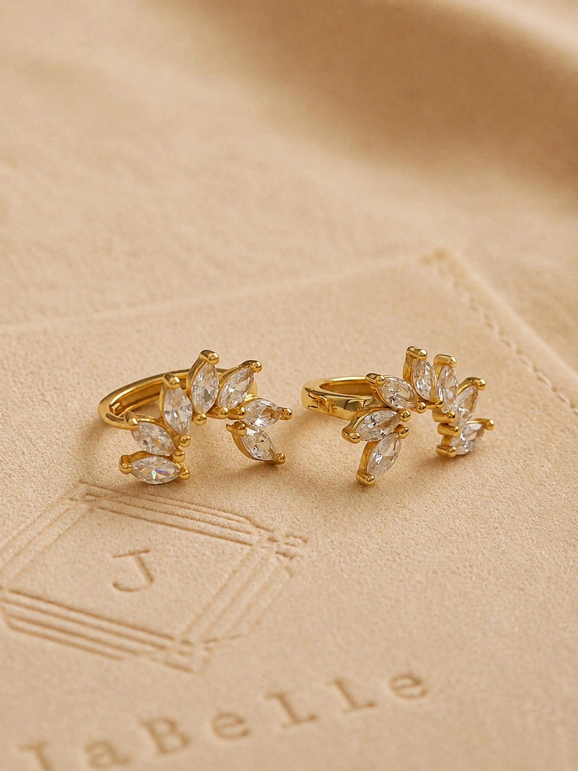 A pair of gold flower huggie earrings featuring floral arrangement shape with cz gemstone, displayed on a luxury beige stone surface.