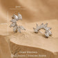 A pair of silver flower huggie earrings featuring floral arrangement shape with cz gemstone, displayed on a luxury beige stone surface.