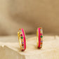 Pair of pink enamel huggie hoop earrings with multicolor cubic zirconia gemstones set in 18k gold plated sterling silver, displayed on a stone background.