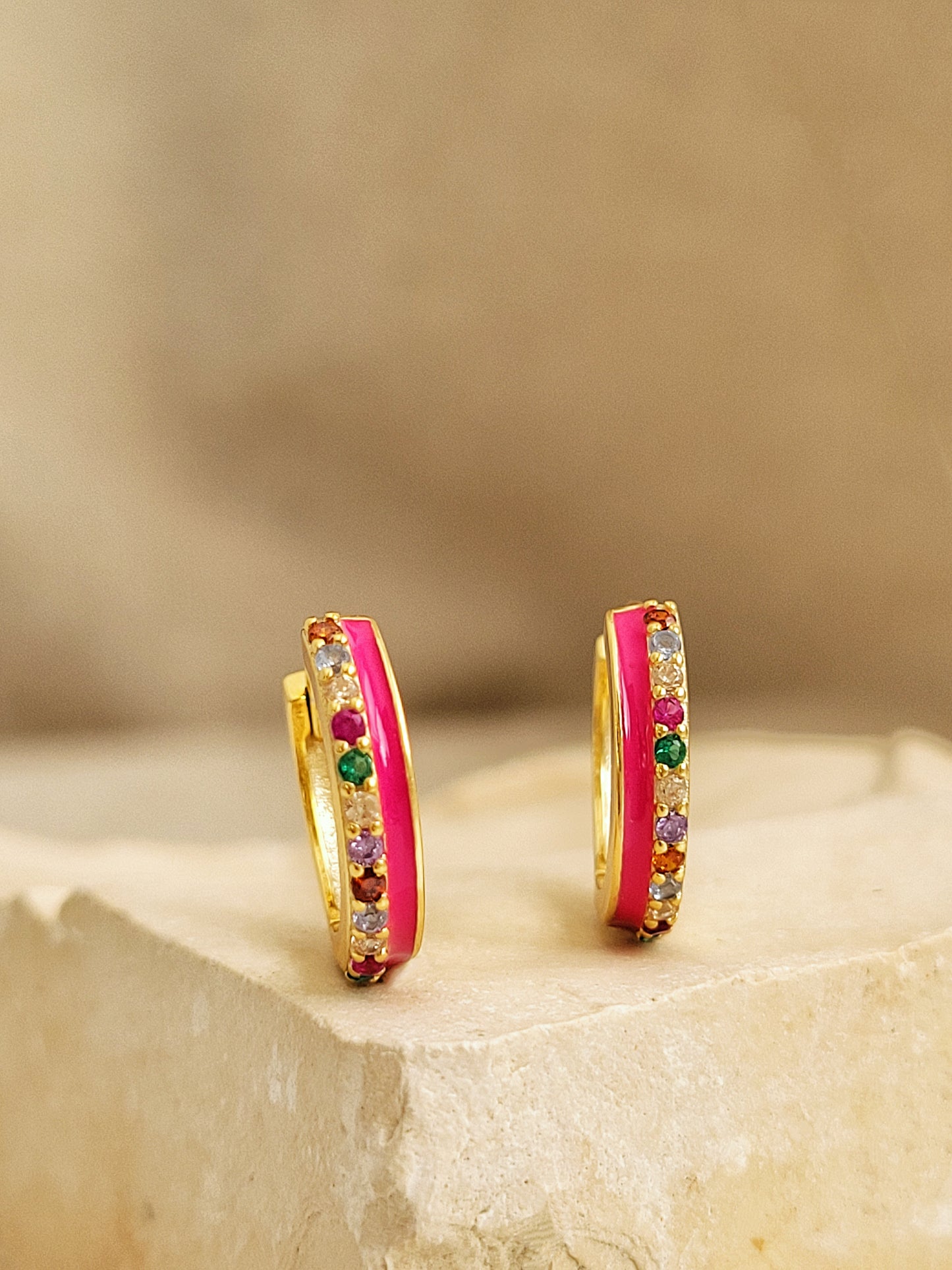 Pair of pink enamel huggie hoop earrings with multicolor cubic zirconia gemstones set in 18k gold plated sterling silver, displayed on a stone background.