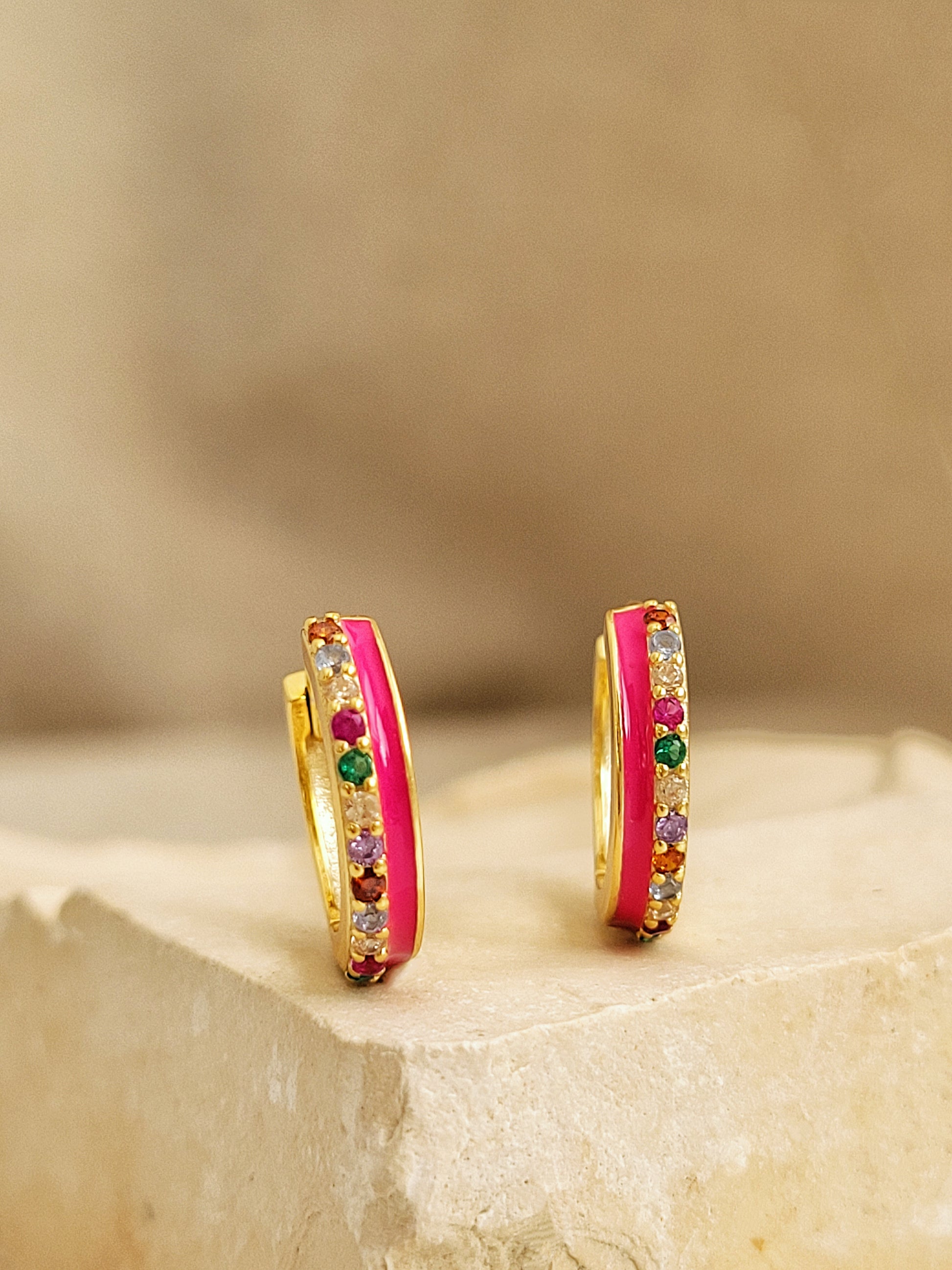 Pair of pink enamel huggie hoop earrings with multicolor cubic zirconia gemstones set in 18k gold plated sterling silver, displayed on a stone background.