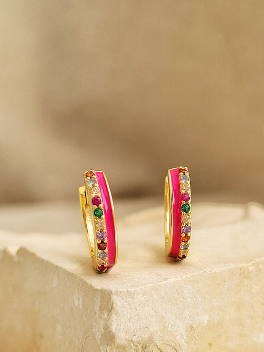 Pair of pink enamel huggie hoop earrings with multicolor cubic zirconia gemstones set in 18k gold plated sterling silver, displayed on a stone background.