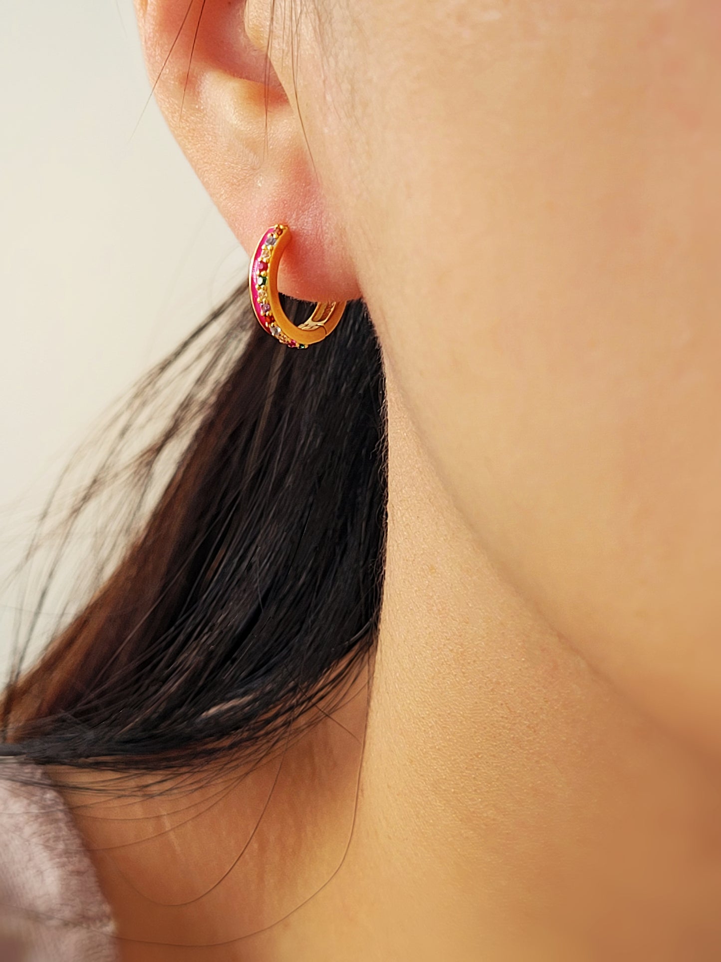 Model wearing pink enamel rainbow huggie hoop earrings with gold finish and multicolor CZ stones, showing small hoop size and elegant fit on ear.