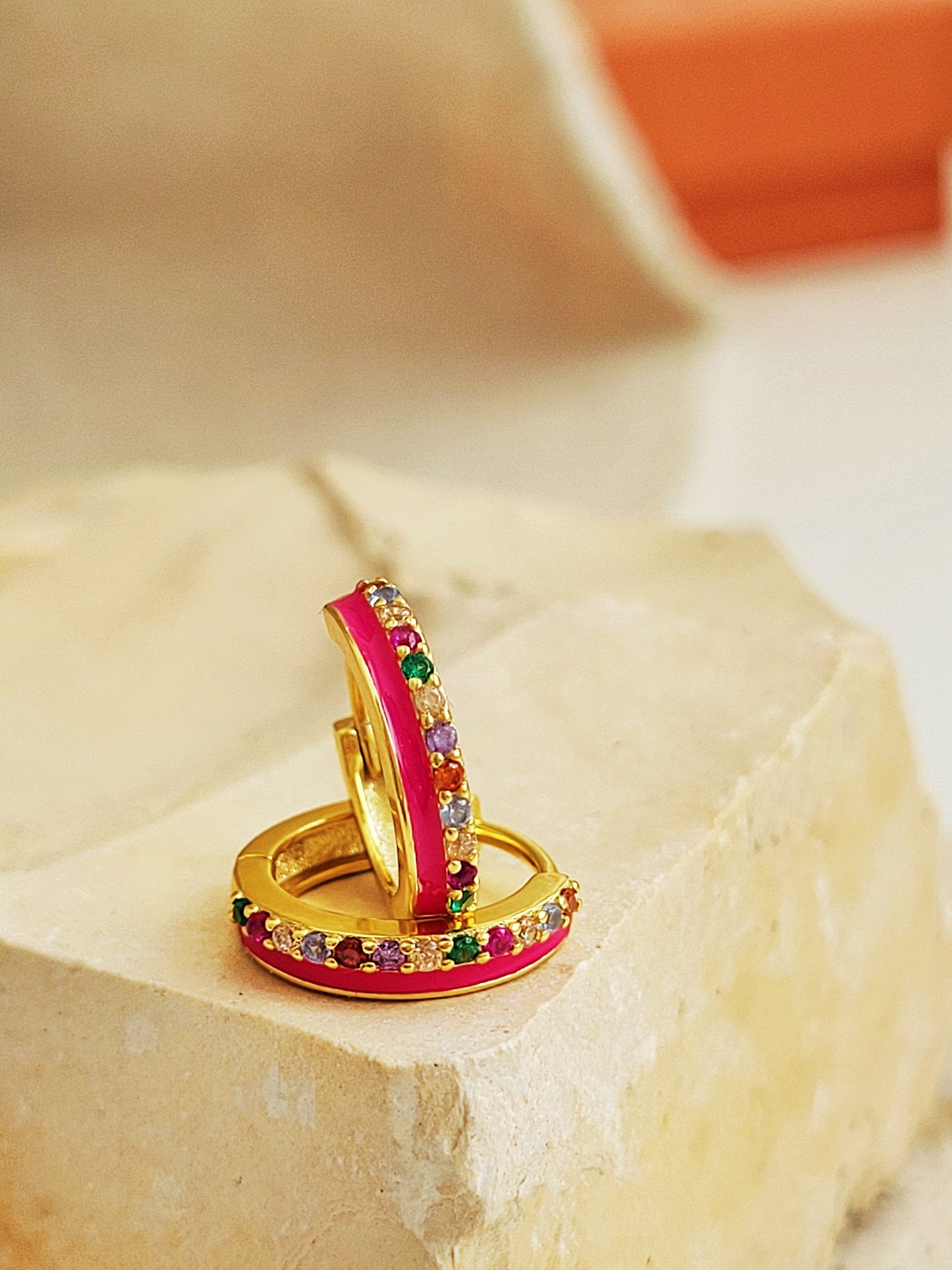 Pair of pink enamel huggie hoop earrings with multicolor cubic zirconia gemstones set in 18k gold plated sterling silver, displayed on a stone background.