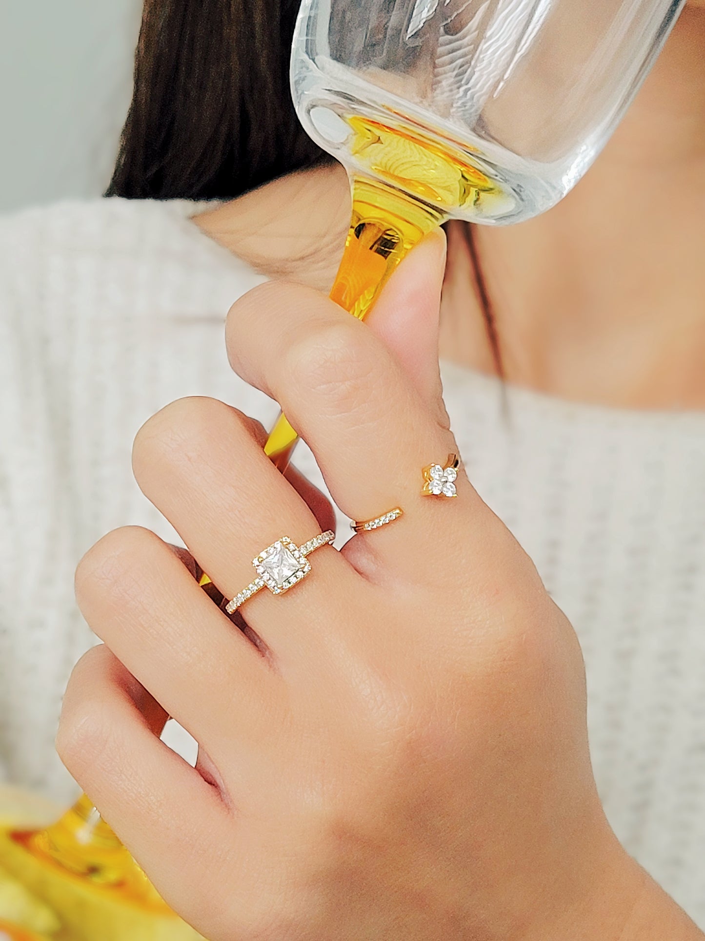 A close-up of a woman's hand holding a champagne glass with an elegant ring showcases a radiant square-cut diamond surrounded by a halo of smaller diamonds, set on a delicate diamond-encrusted gold band.