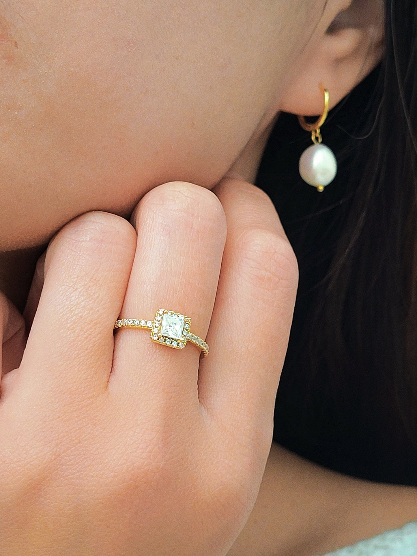 A close-up of a woman's hand adorned with an elegant ring showcases a radiant square-cut diamond surrounded by a halo of smaller diamonds, set on a delicate diamond-encrusted gold band.