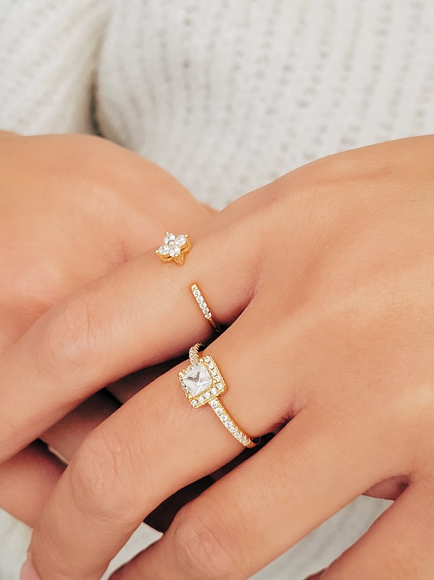 A close-up of a woman's hand adorned with elegant rings. One ring showcases a radiant princess cut diamond surrounded by a halo of smaller diamonds, set on a delicate diamond-encrusted gold band.