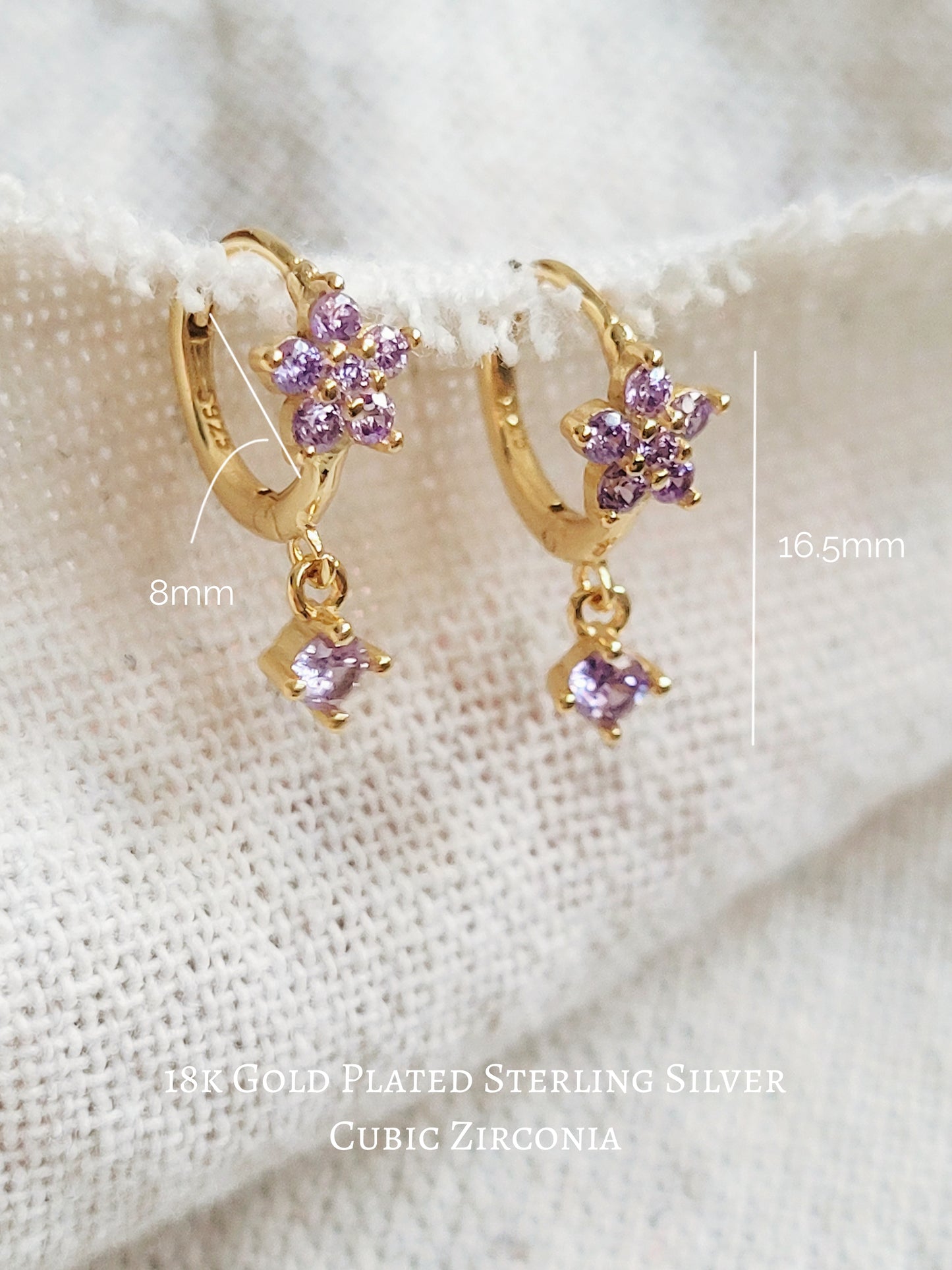 Close-up of a pair of dainty purple flower huggie earrings with dangling charm in 18k gold plated sterling silver and cubic zirconia.