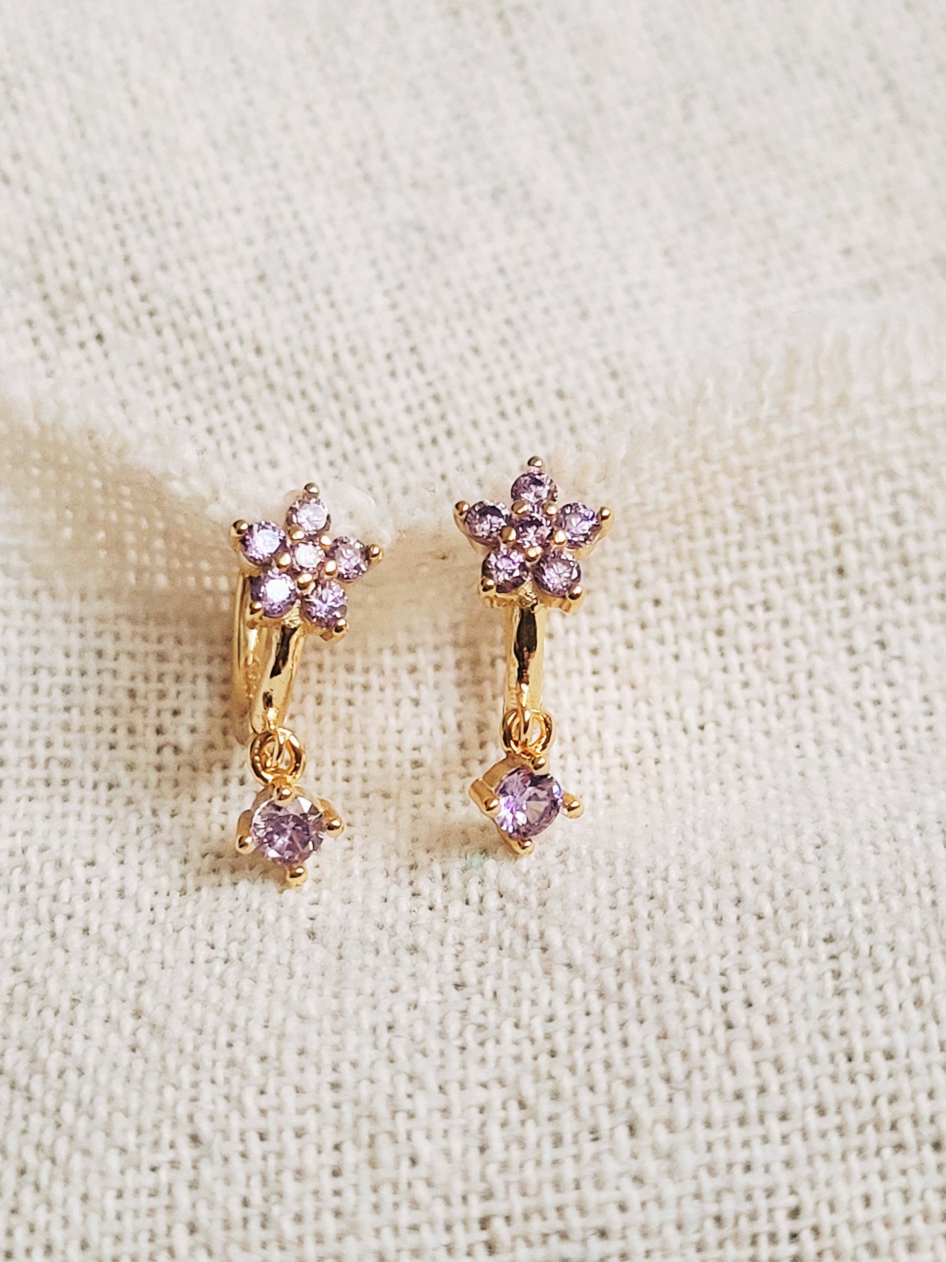 Close-up of a pair of dainty purple flower huggie earrings with dangling charm in 18k gold plated sterling silver and cubic zirconia.