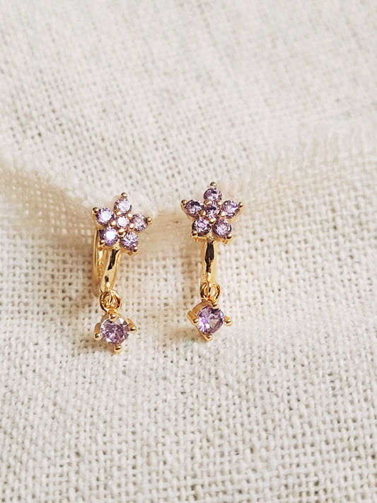 Close-up of a pair of dainty purple flower huggie earrings with dangling charm in 18k gold plated sterling silver and cubic zirconia.