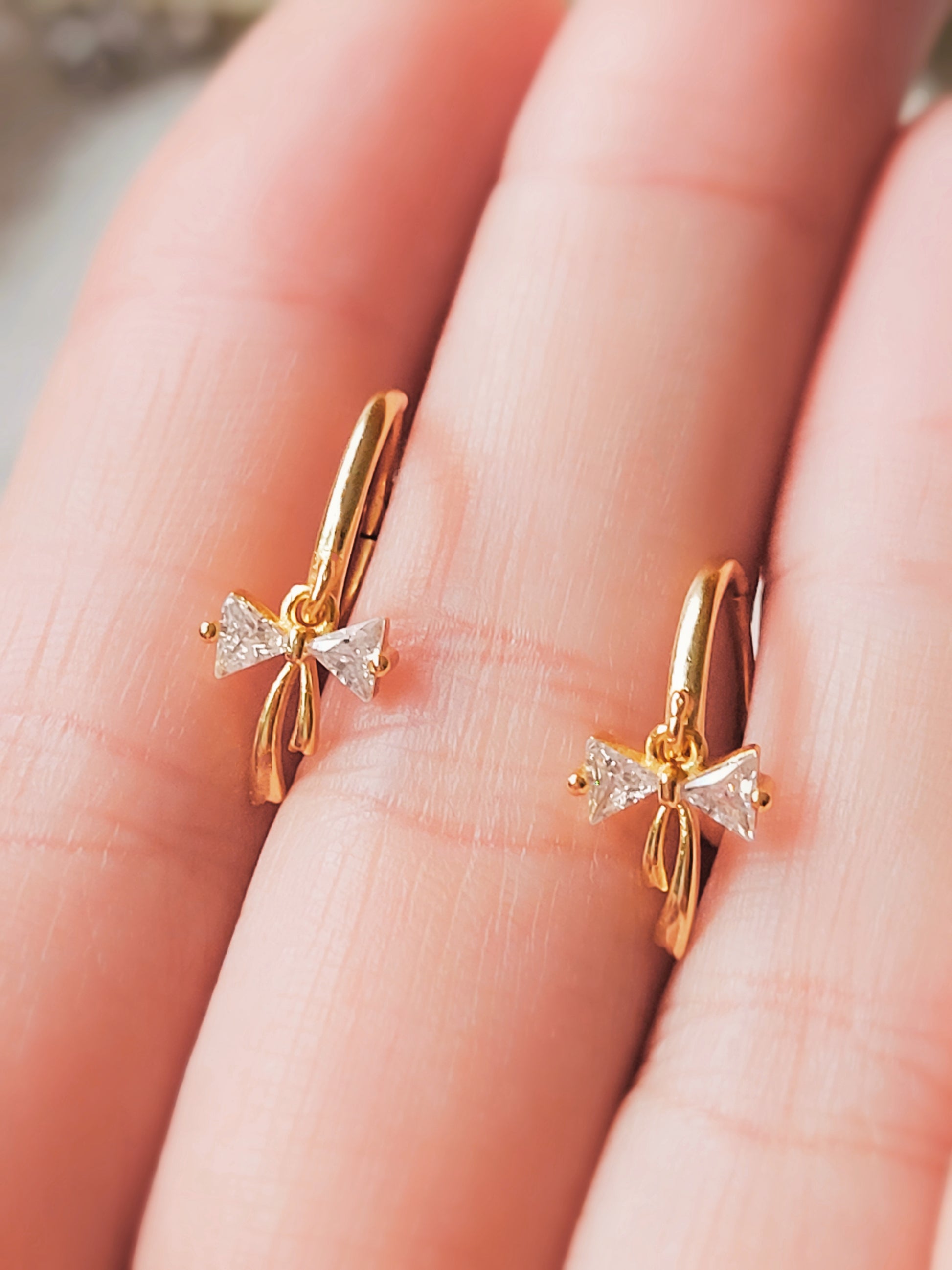 A pair of dainty gold plated ribbon bow dangly huggie earrings displayed on a woman&#39;s hand.