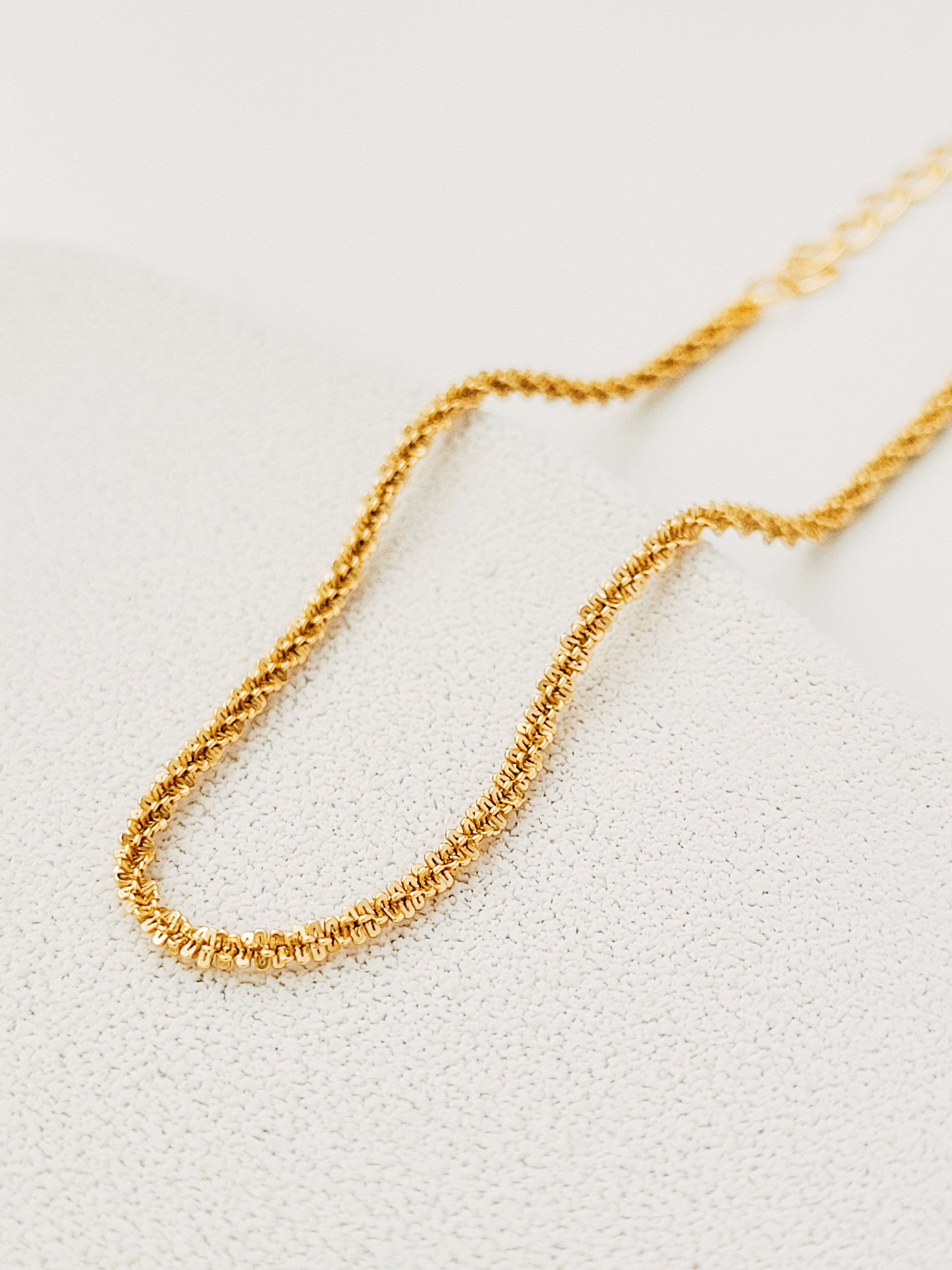 Gold Rope chain bracelet with intricate, small interlocking links, laying in a gentle curve on a beige textured background, creating a sophisticated look.