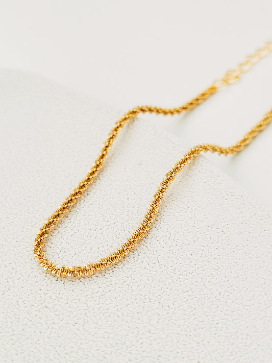 Gold Rope chain bracelet with intricate, small interlocking links, laying in a gentle curve on a beige textured background, creating a sophisticated look.