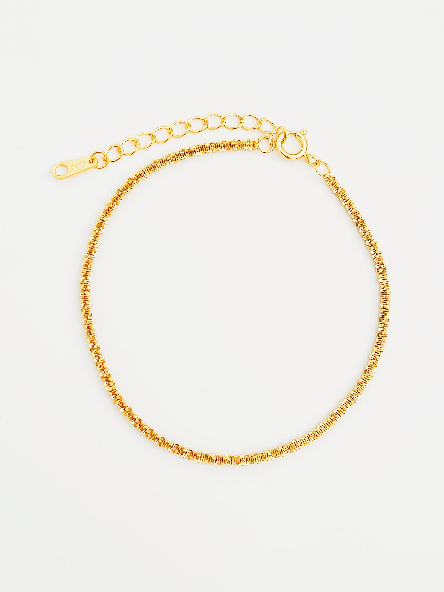 Detailed view of a gold rope chain bracelet with tightly woven links, arranged against a light, textured surface that highlights its fine craftsmanship.