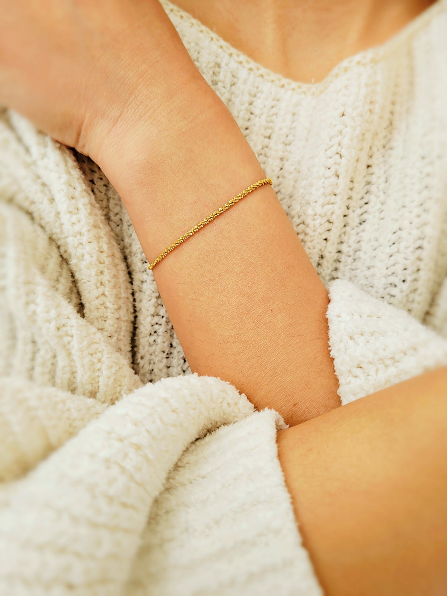 A woman’s arm adorned with a minimalist fine gold rope bracelet, held near her face in a relaxed pose, with hints of a soft, white sweater visible in the background.