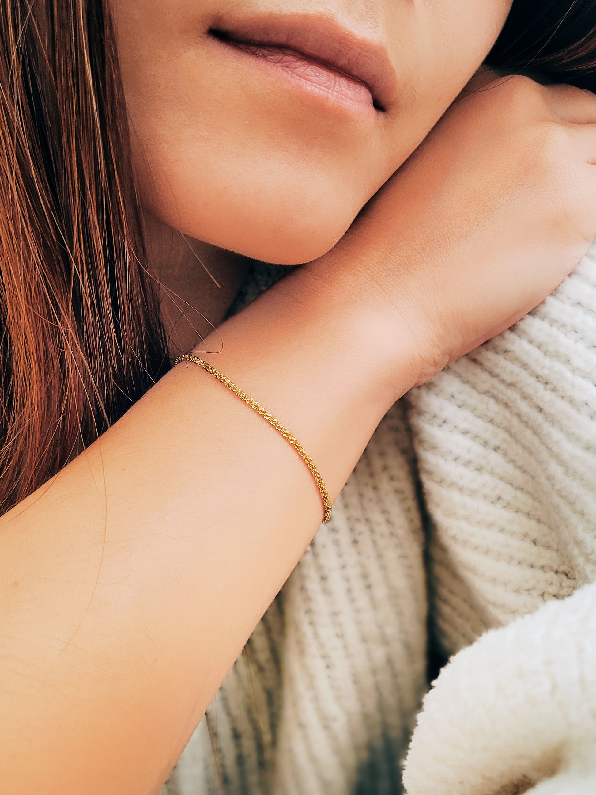 Close-up of a woman resting her arm on her body, wearing a delicate, shimmering gold rope bracelet against her wrist, with a cozy, knit sweater in the background.