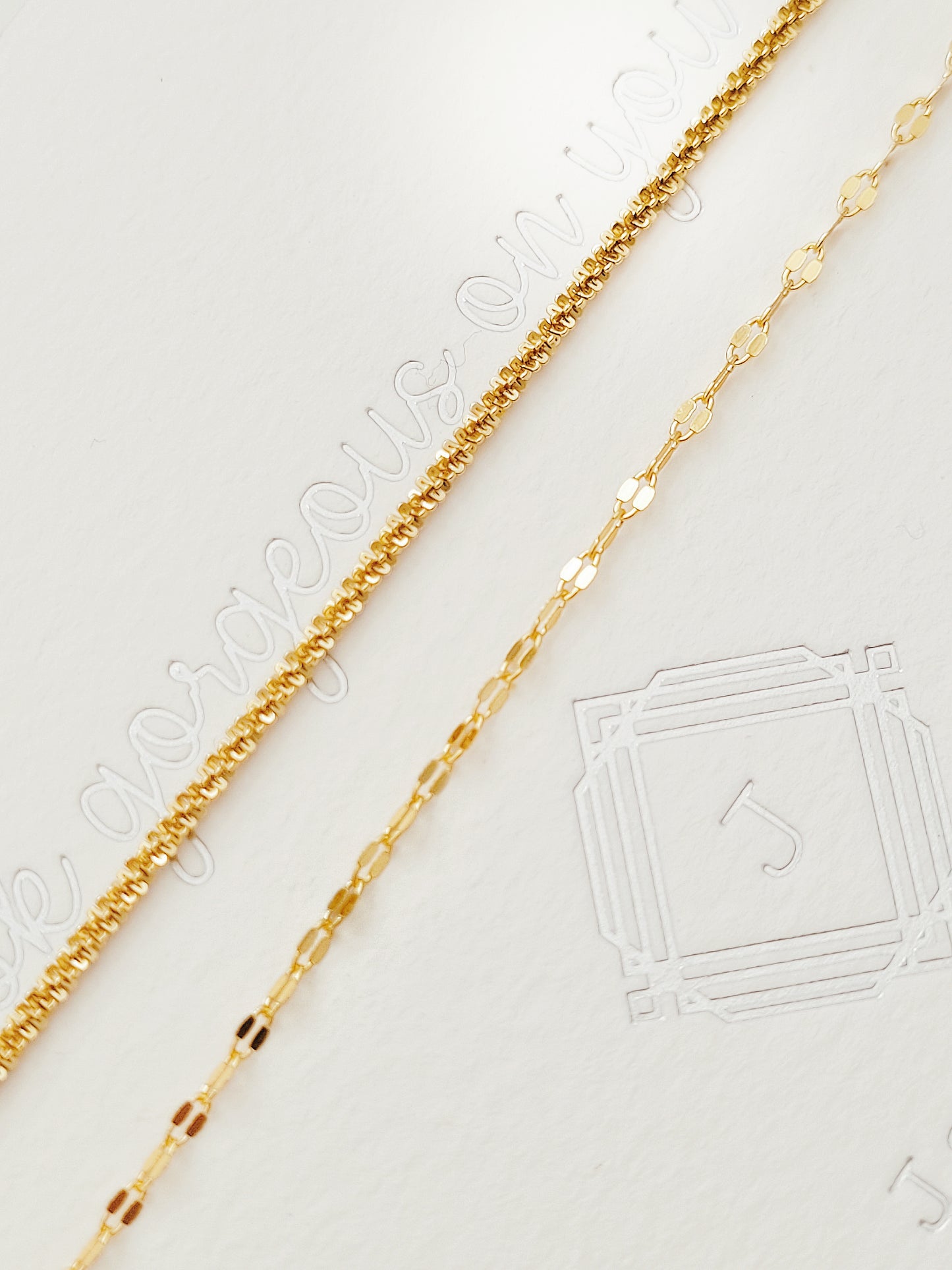 Two textured delicate and simple gold chain bracelets elegantly draped on a light beige, subtly textured background, showcasing its intricate design.