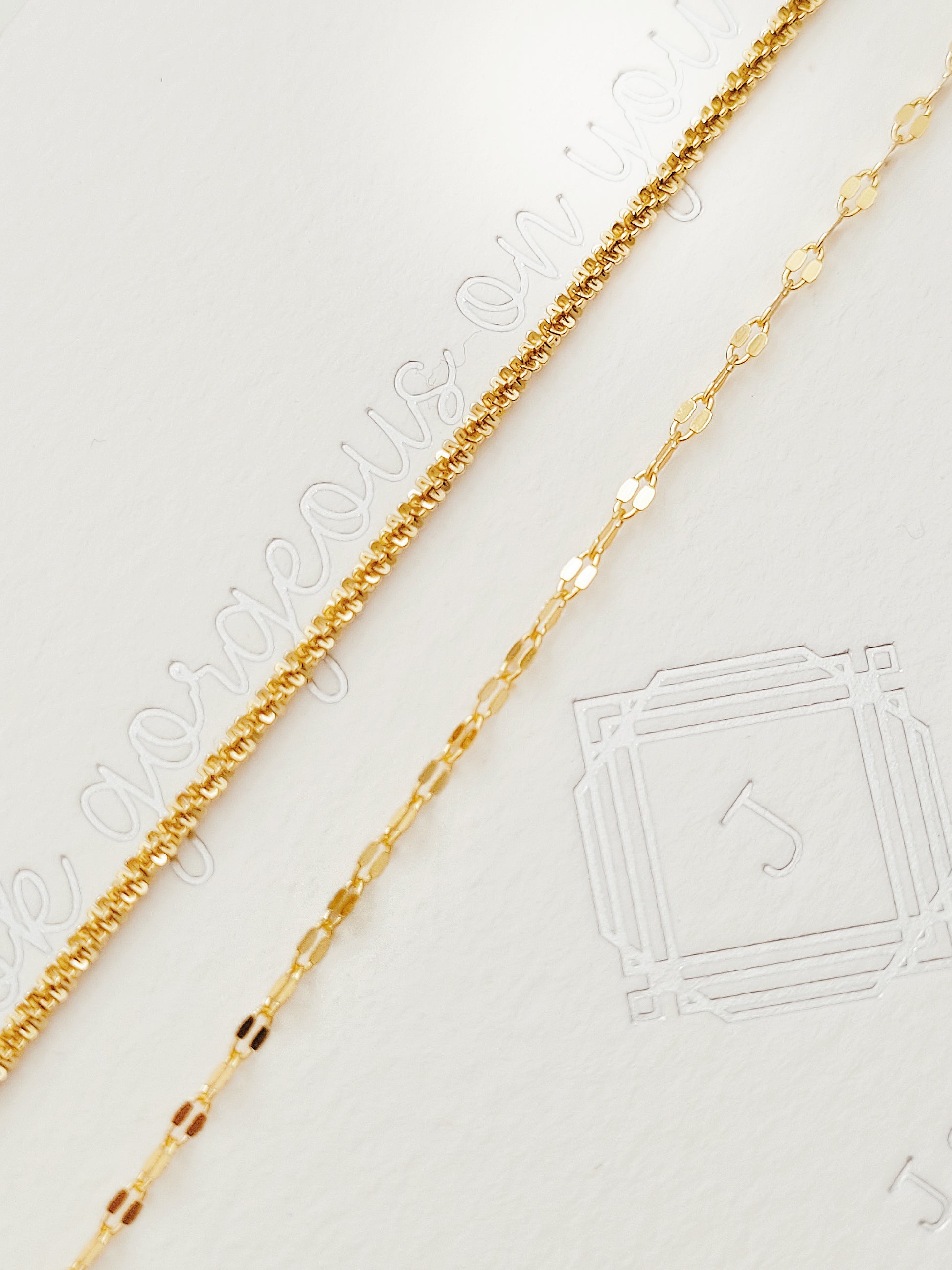 Two textured delicate and simple gold chain bracelets elegantly draped on a light beige, subtly textured background, showcasing its intricate design.