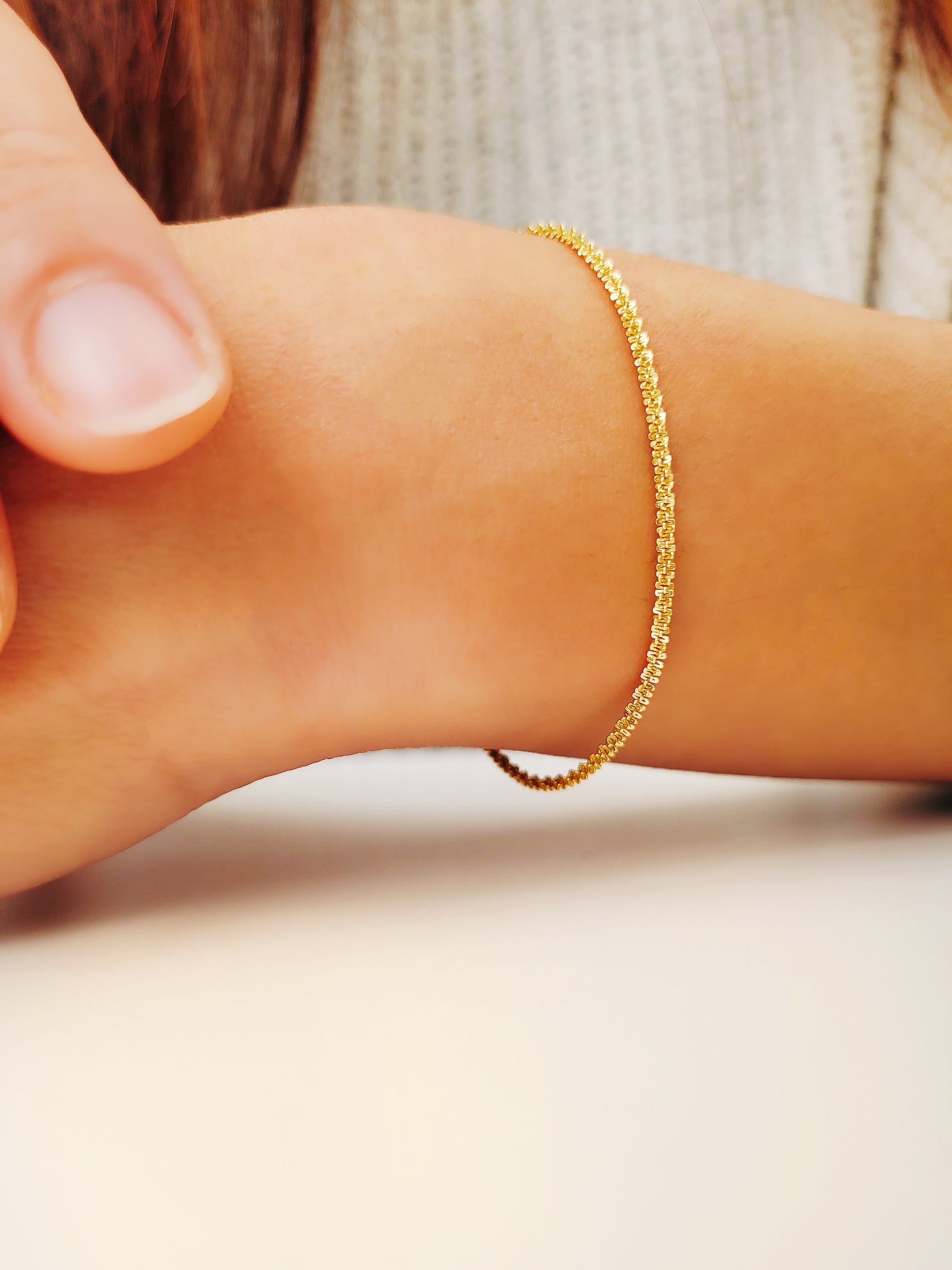 A woman’s arm adorned with a dainty fine gold rope bracelet, held by another hand.