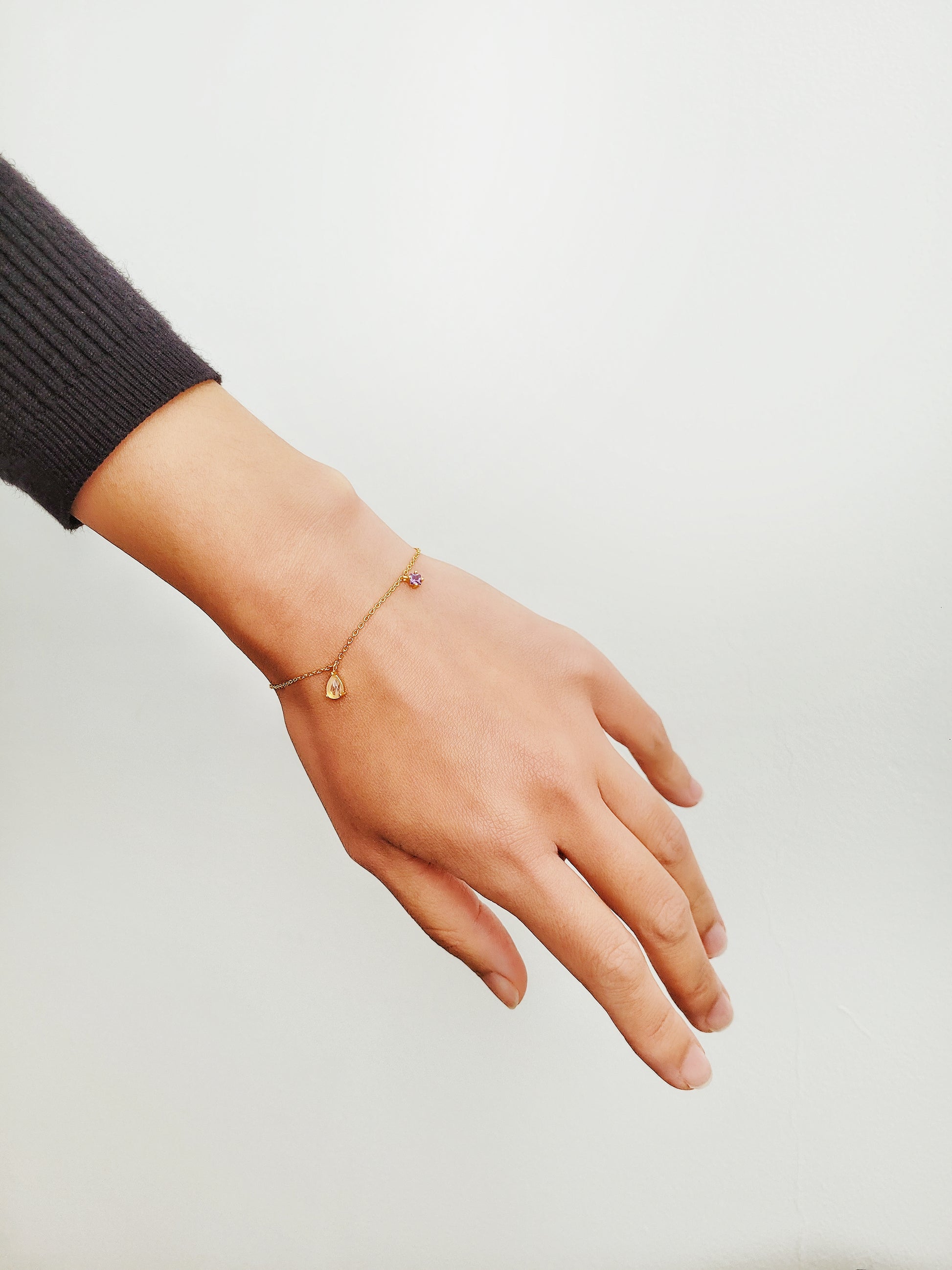 A woman's hand displays a luxurious gold plated bracelet featuring a captivating gold teardrop Rutilated Quartz charm and an exquisite purple Amethyst charm.
